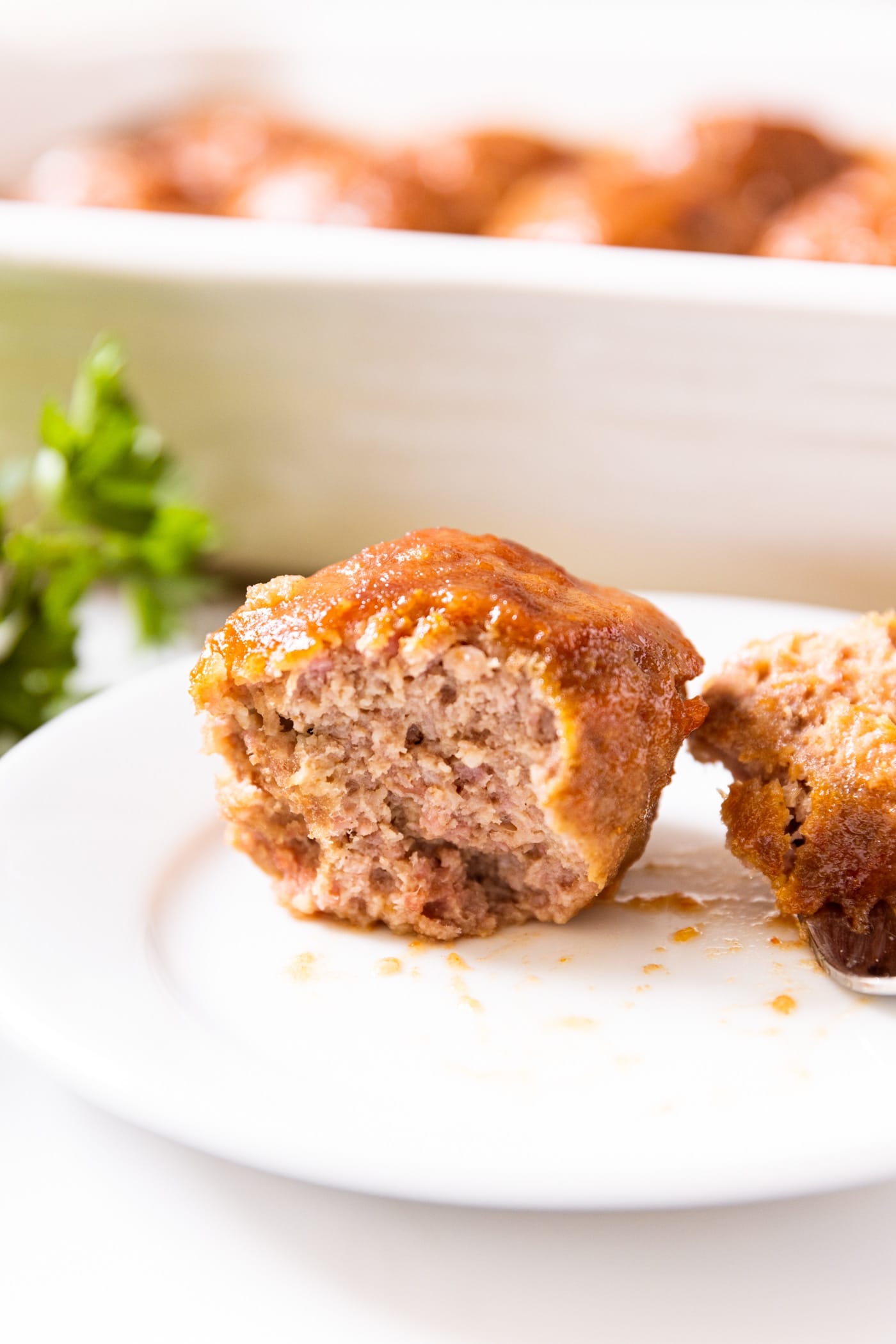 Side profile of ham ball sliced in half with fork holding piece off to side with baking dish in background filled with more ham balls
