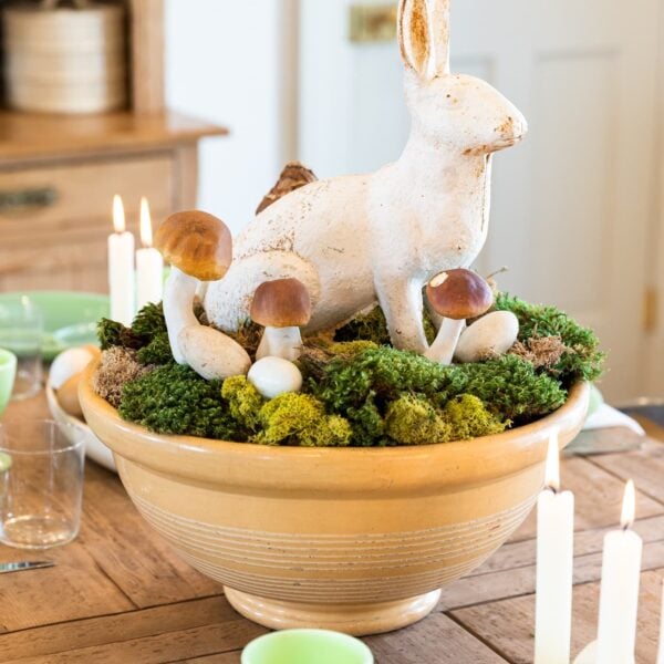 White metal rabbit sitting among moss, eggs, and mushrooms all in a yellow bowl sitting on wood dining table