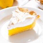 White plate with slice of lemon meringue pie with white meringue topping with rest of pie in background