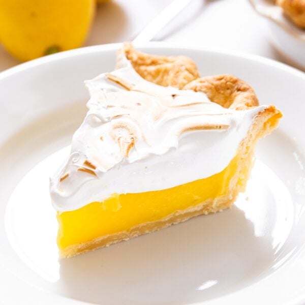 White plate with slice of lemon meringue pie with white meringue topping with rest of pie in background