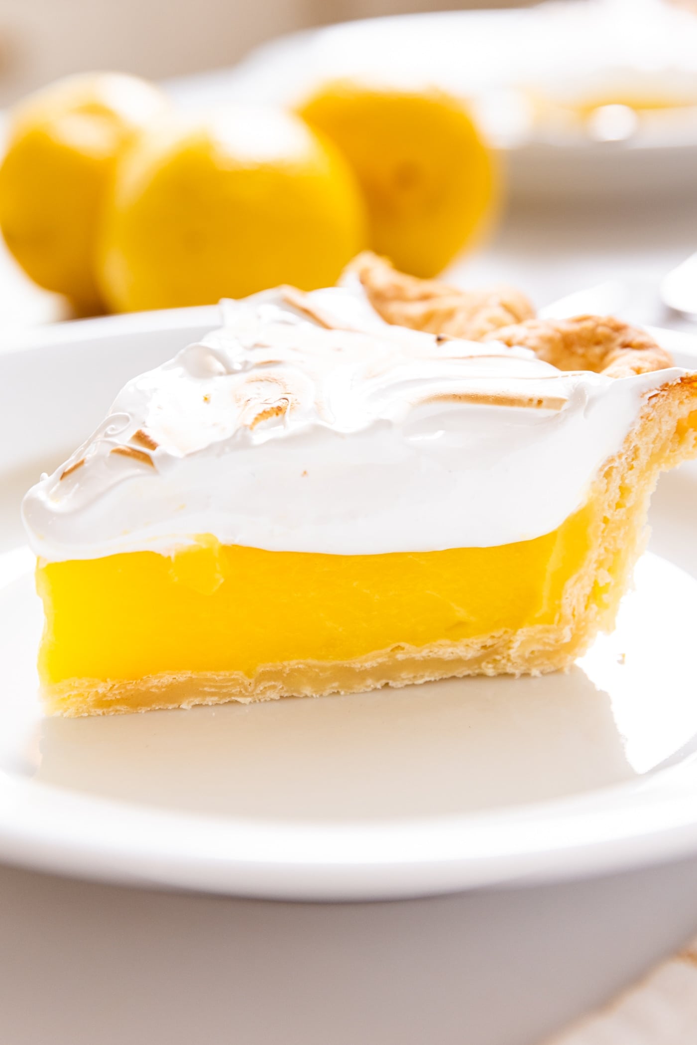 Side view of slice of lemon meringue pie sitting on white plate with lemons in background as well as other pie plate