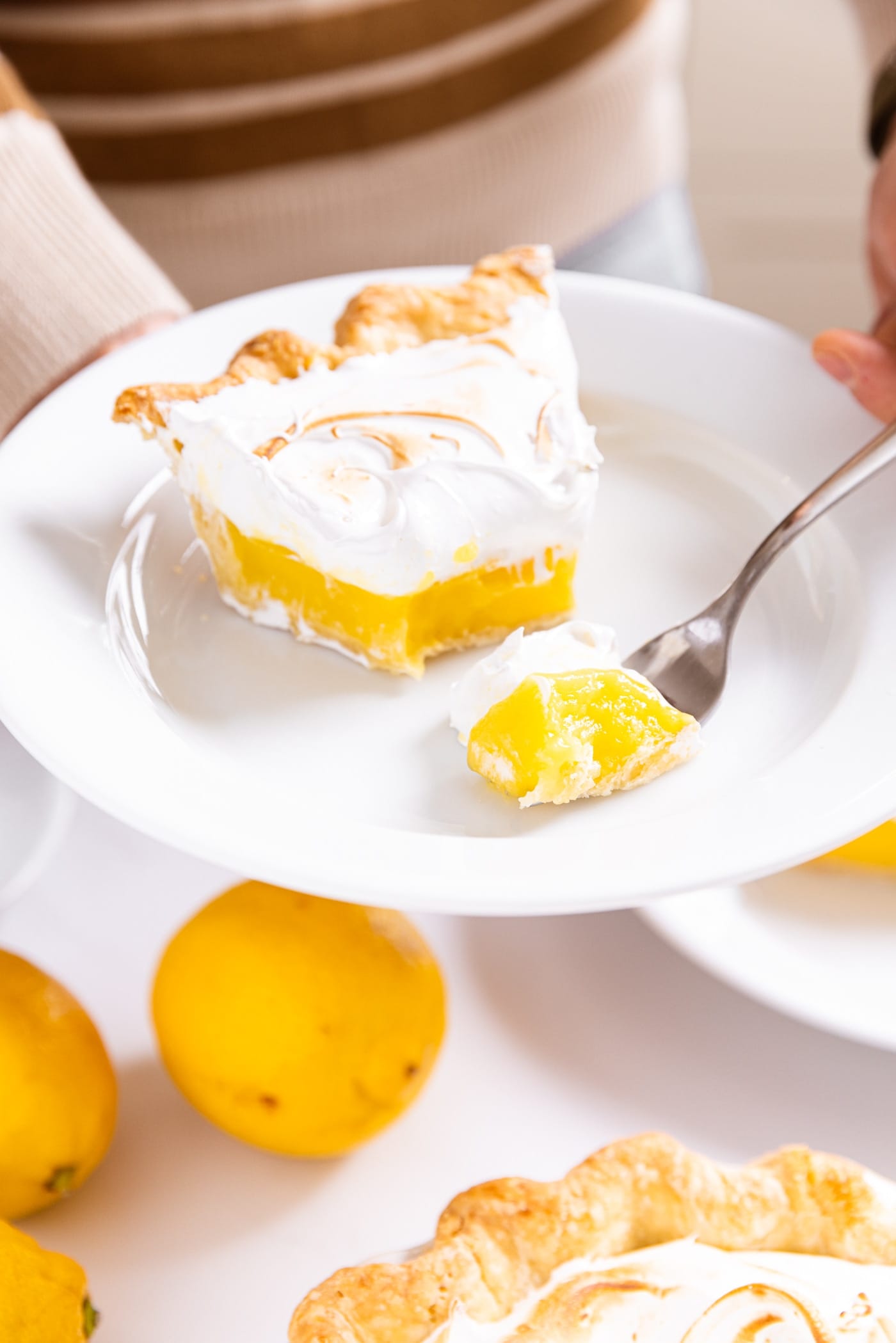 Hand holding white plate with slice of lemon meringue pie with piece sliced off of tip of pie with more lemons underneath