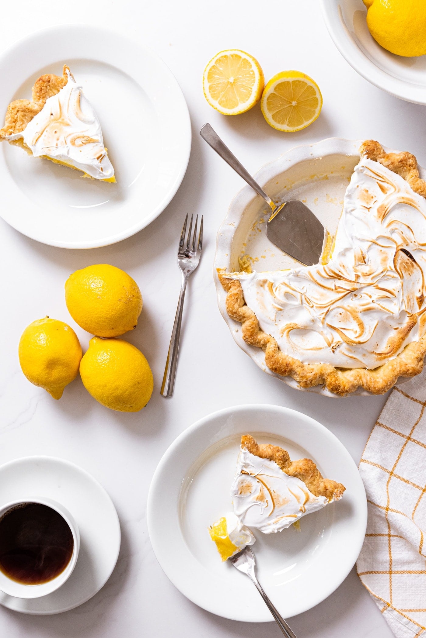 Top down view of white countertop with lemon meringue pie with slices cut and on white plates with other lemons and cup of coffee as well