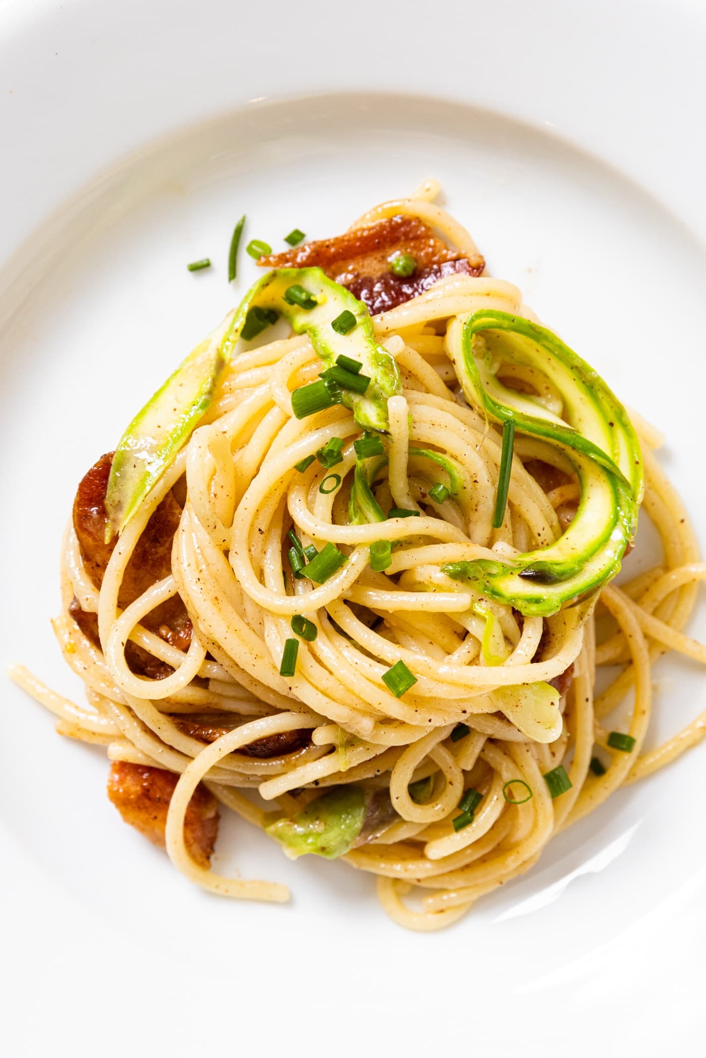 swirled spaghetti sitting on white plate with brown butter sauce on top along with bacon and shaved asparagus.