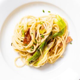 White plate filled with spaghetti noodles with brown butter sauce on top with shaved asparagus and bacon and chive pieces