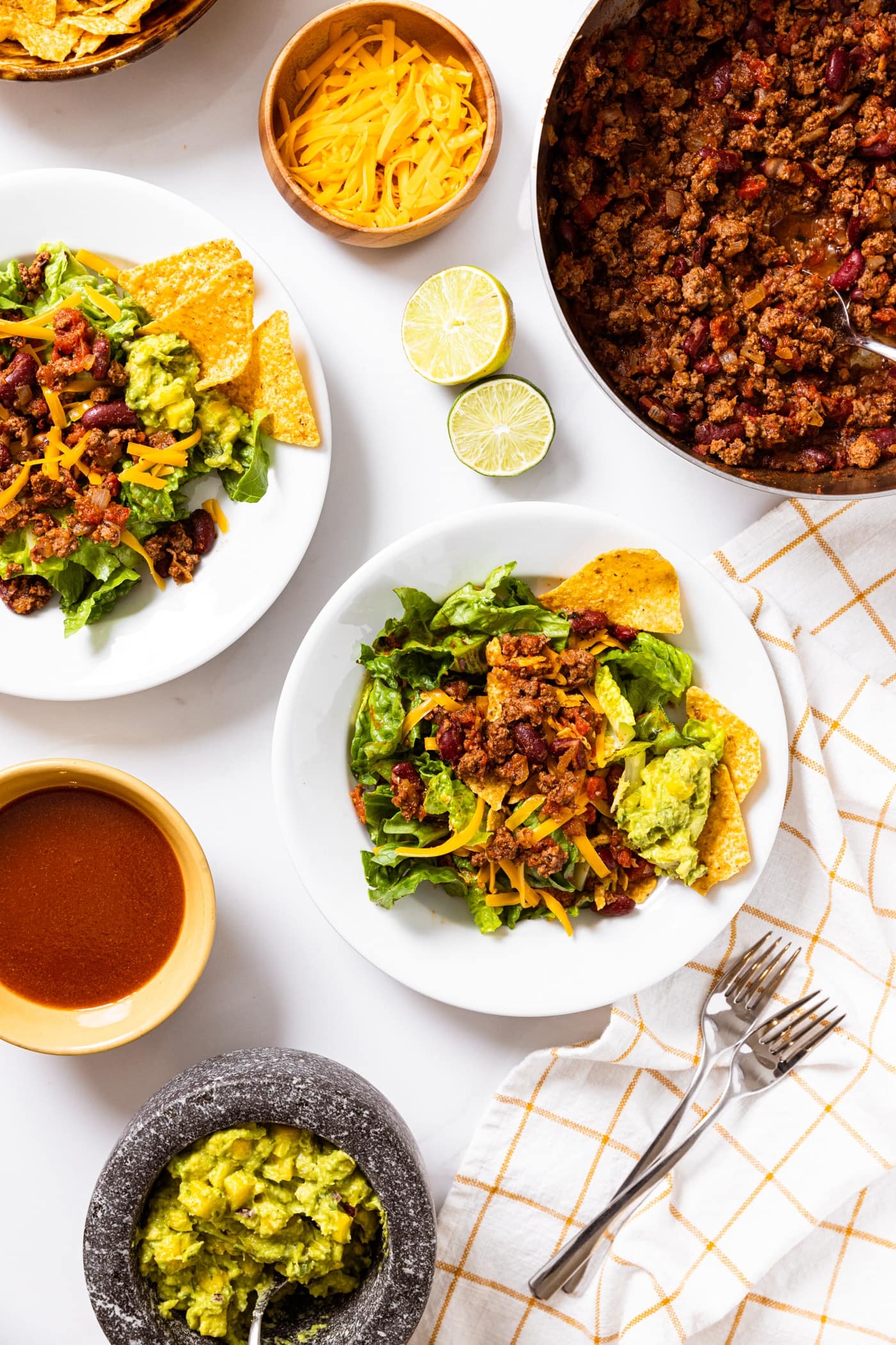 Top down view of taco salad being assembled on plates on white countertop with bowl of meat mixture, plates already filled with taco salad, cheese, limes, red dressing, guacamole, and tortilla chips