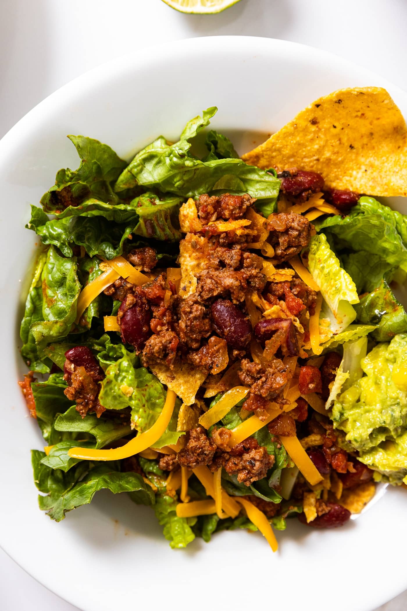 Top down view of white plate filled with green lettuce and topped with hamburger meat making a taco salad with tortilla chips and guacamole on the side