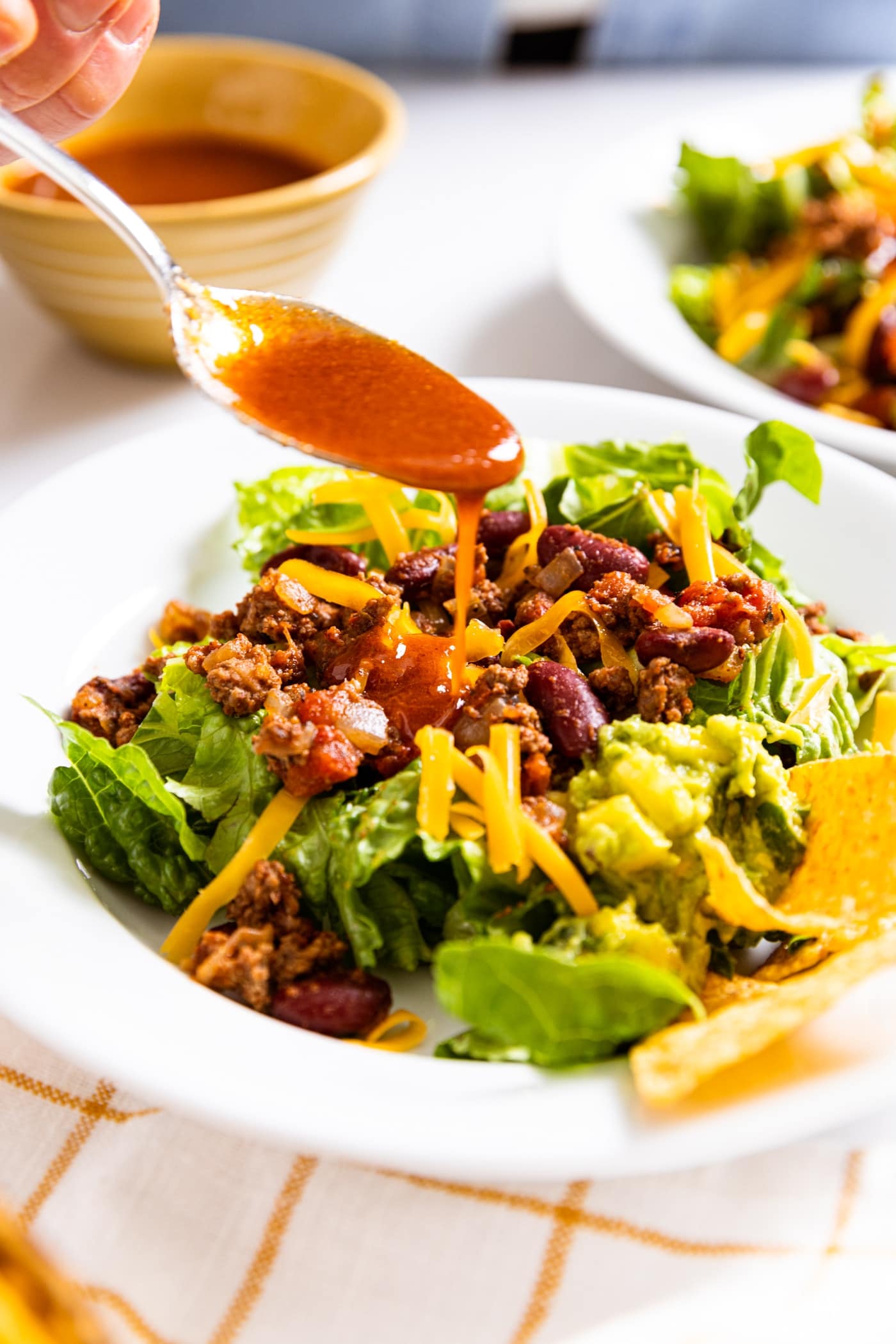 Spoon drizzling red colored salad dressing on top of taco salad with guacamole and tortilla chips on side with container of salad dressing in background