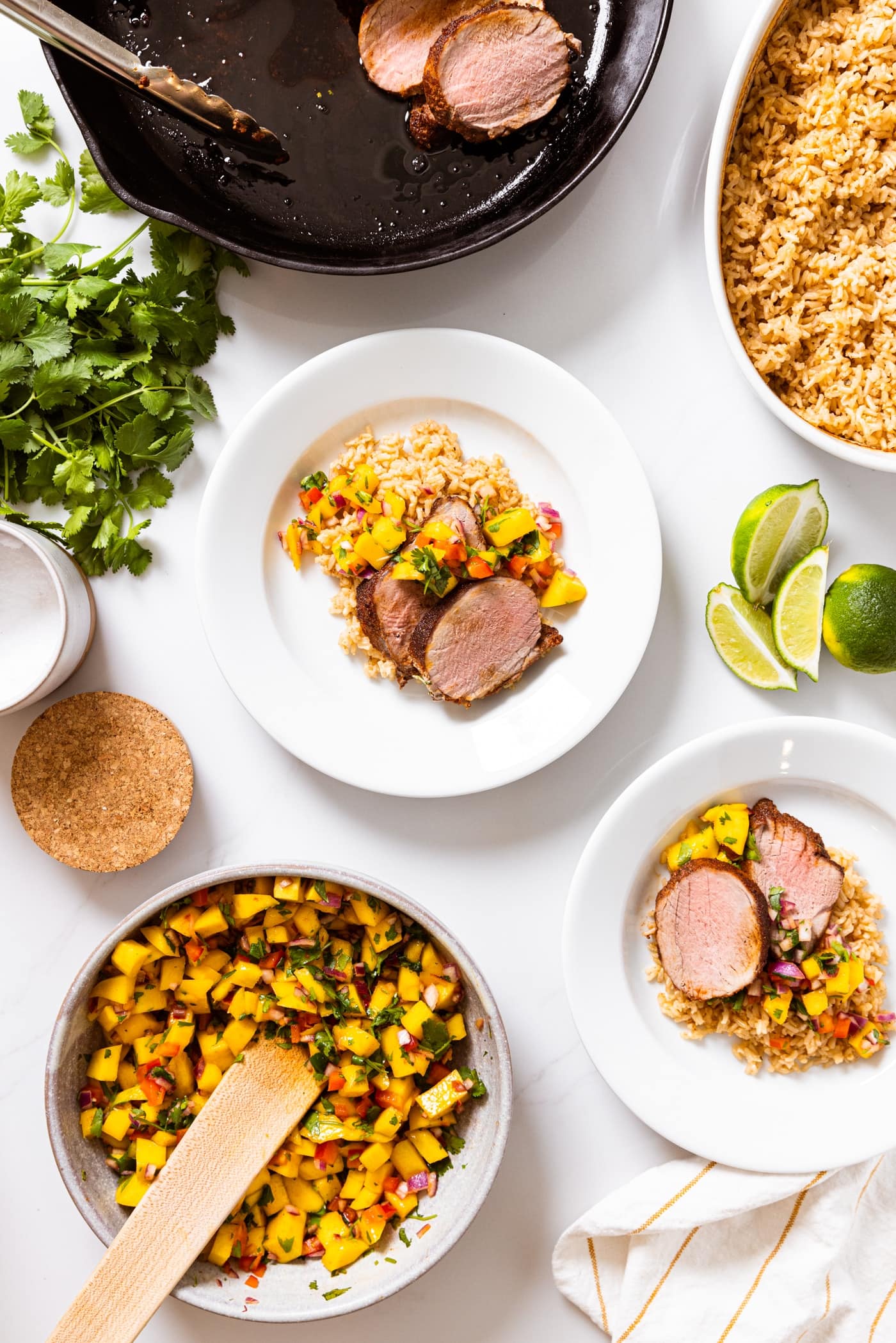 Top down view of white plate filled with Jamaican-style pork tenderloin with other bowls all around containing mango salsa and rice and leftover pieces of tenderloin
