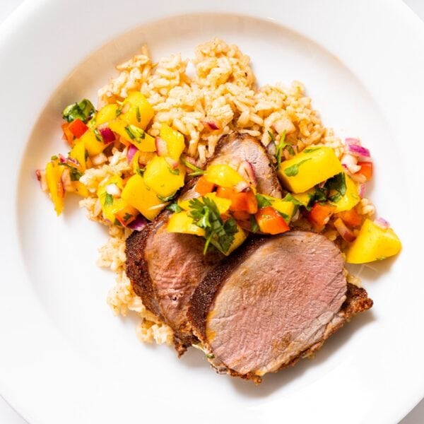 Top down view of white plate filled with slices of pork tenderloin with Jamaican-style crust with brown rice underneath with yellow colored mango salsa all over the top