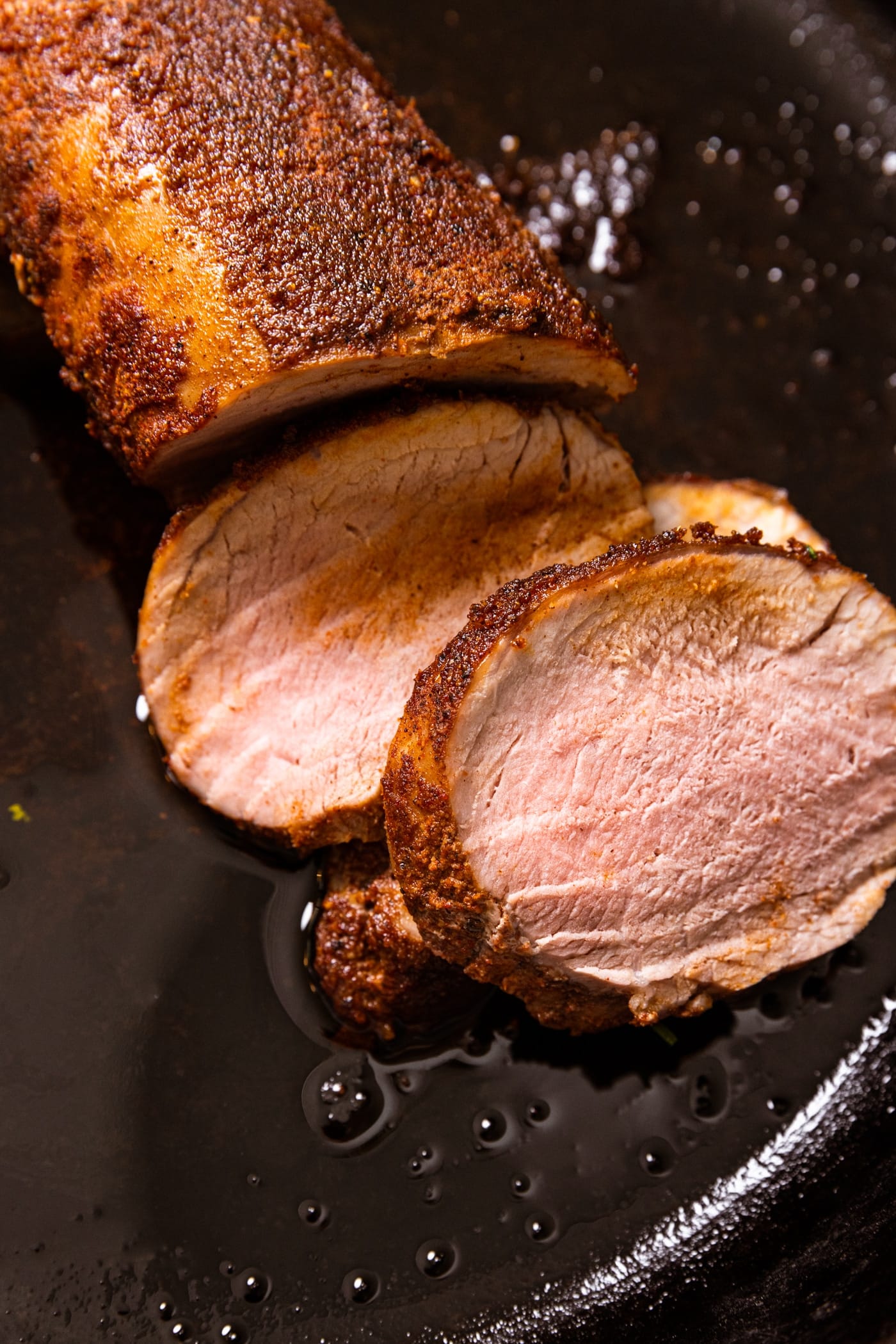 Slices of pork tenderloin with Jamaican-style spices on top sitting in black cast iron skillet with extra oil