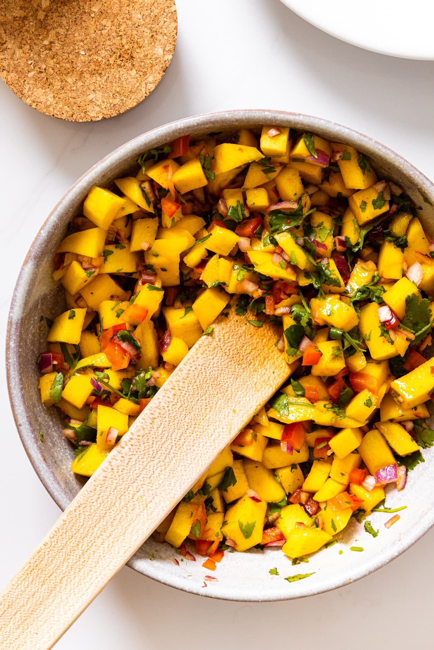 Top down view of ceramic bowl filled with orange colored mango salsa with bits of red pepper and cilantro with wooden spoon in bowl