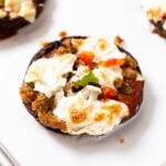 Round portobello mushroom sitting on white piece of parchment paper on baking sheet topped with sausage, red and green pepper, and melted cheese