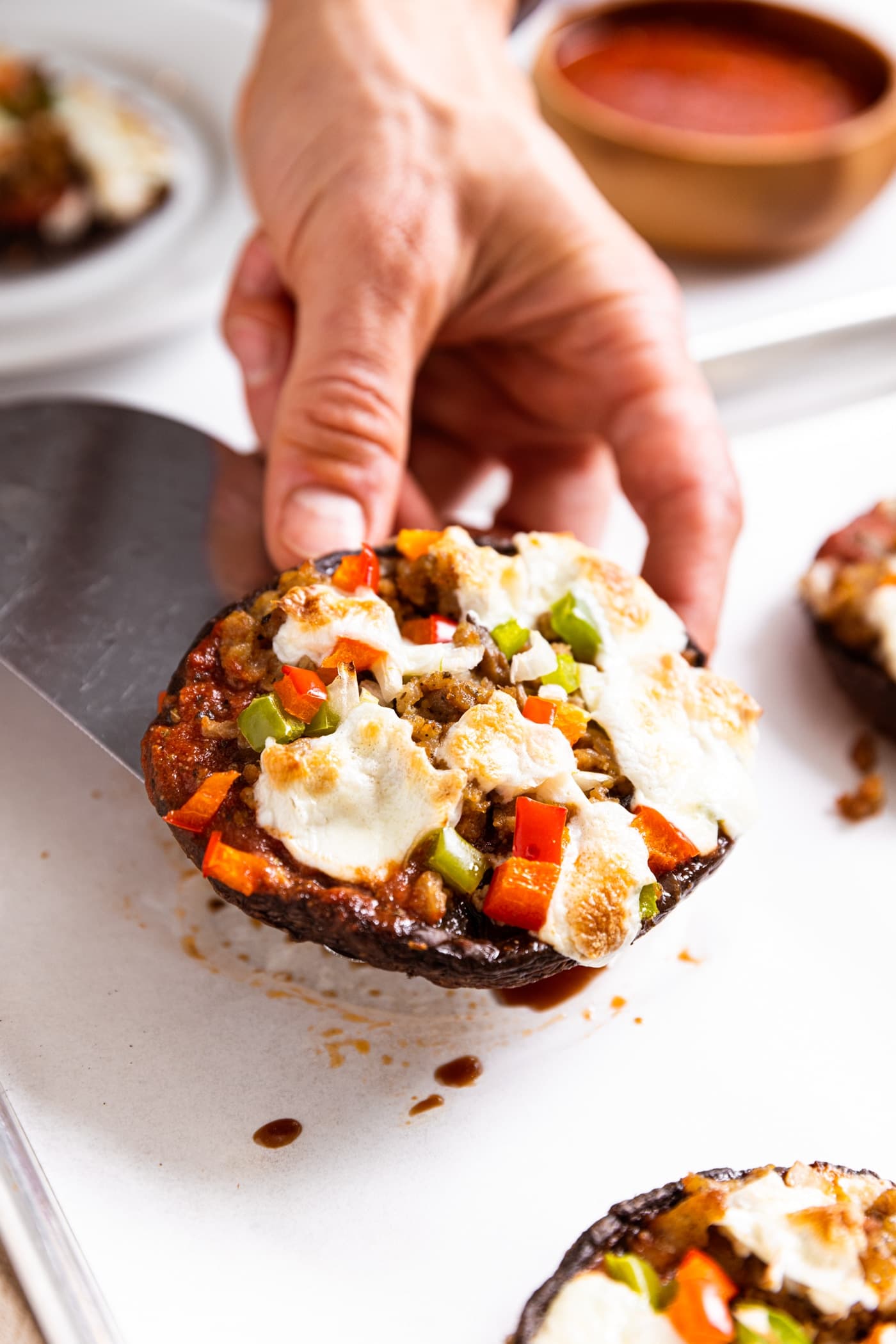 Hand holding metal spatula with portobello mushroom pizza with cheese, red and green pepper, and sausage on top