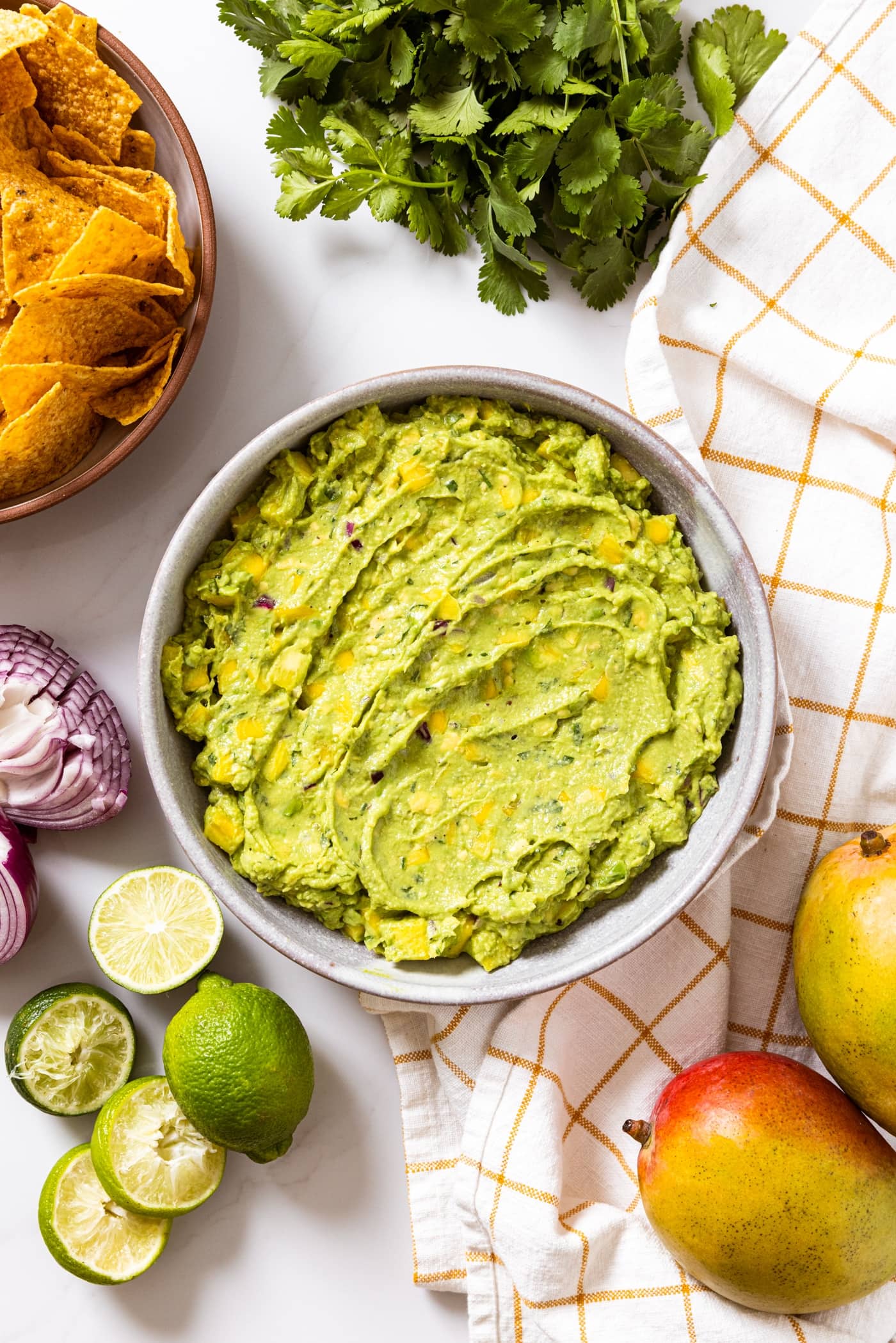 Top down view of white bowl filled with green colored tropical guacamole sitting on white countertop with squeezed limes, red onion, cilantro, mango, and tortilla chips all around
