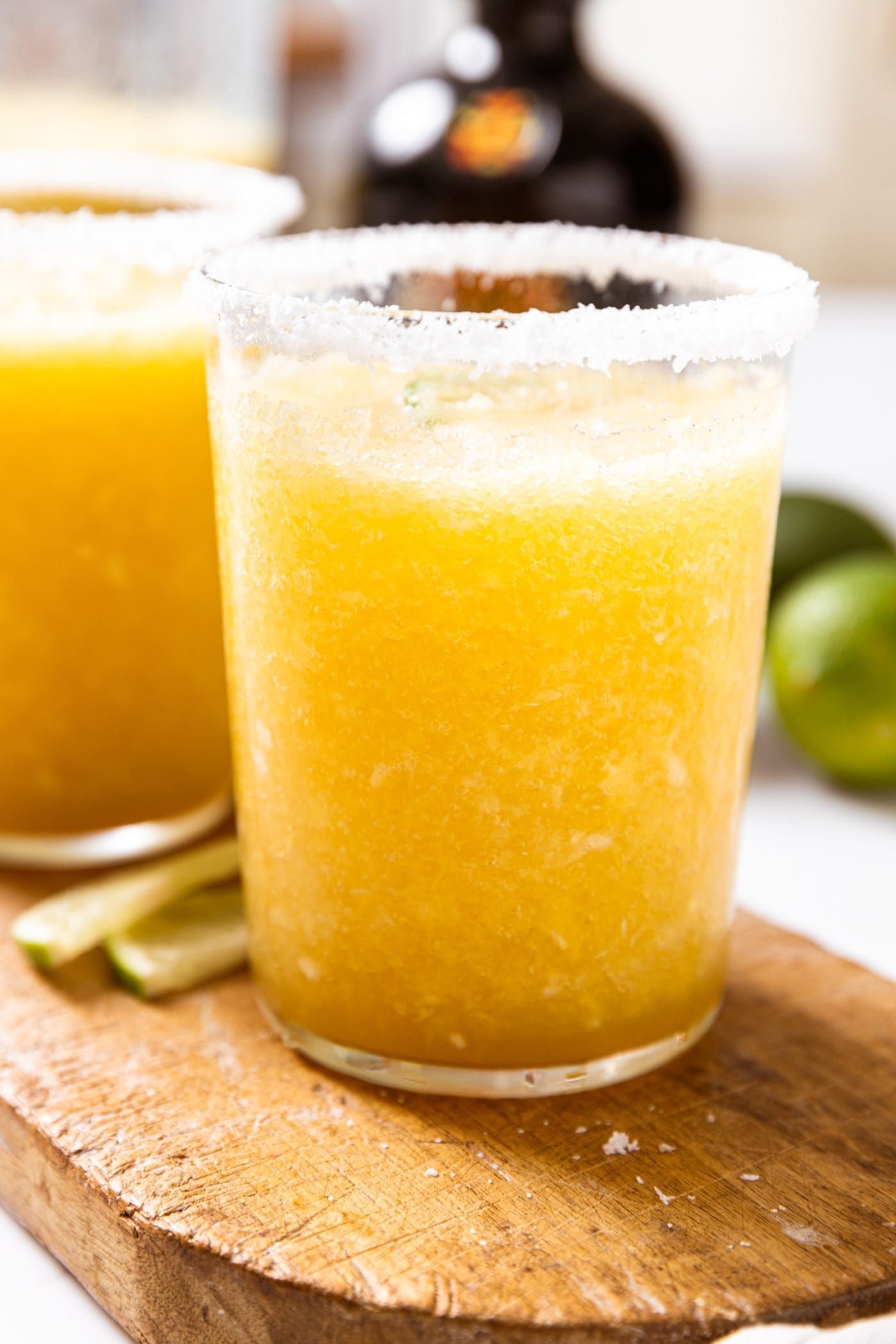 Large glass filled with yellow colored blended margarita sitting on wood board with glass rimmed with salt and lime slices to the side