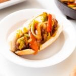 White plate filled with beer brat in hot dog bun with sautéed peppers on top all on white surface.
