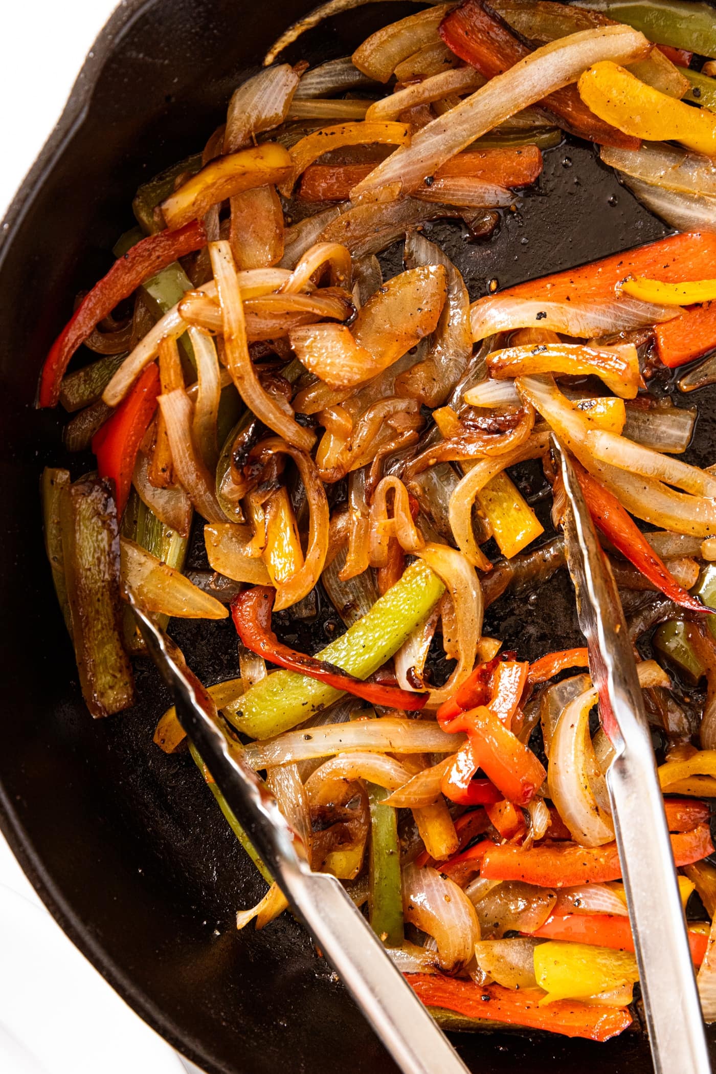 Black cast iron skillet sitting on white surface filled with sautéed bell peppers and onion with tongs resting in pan.