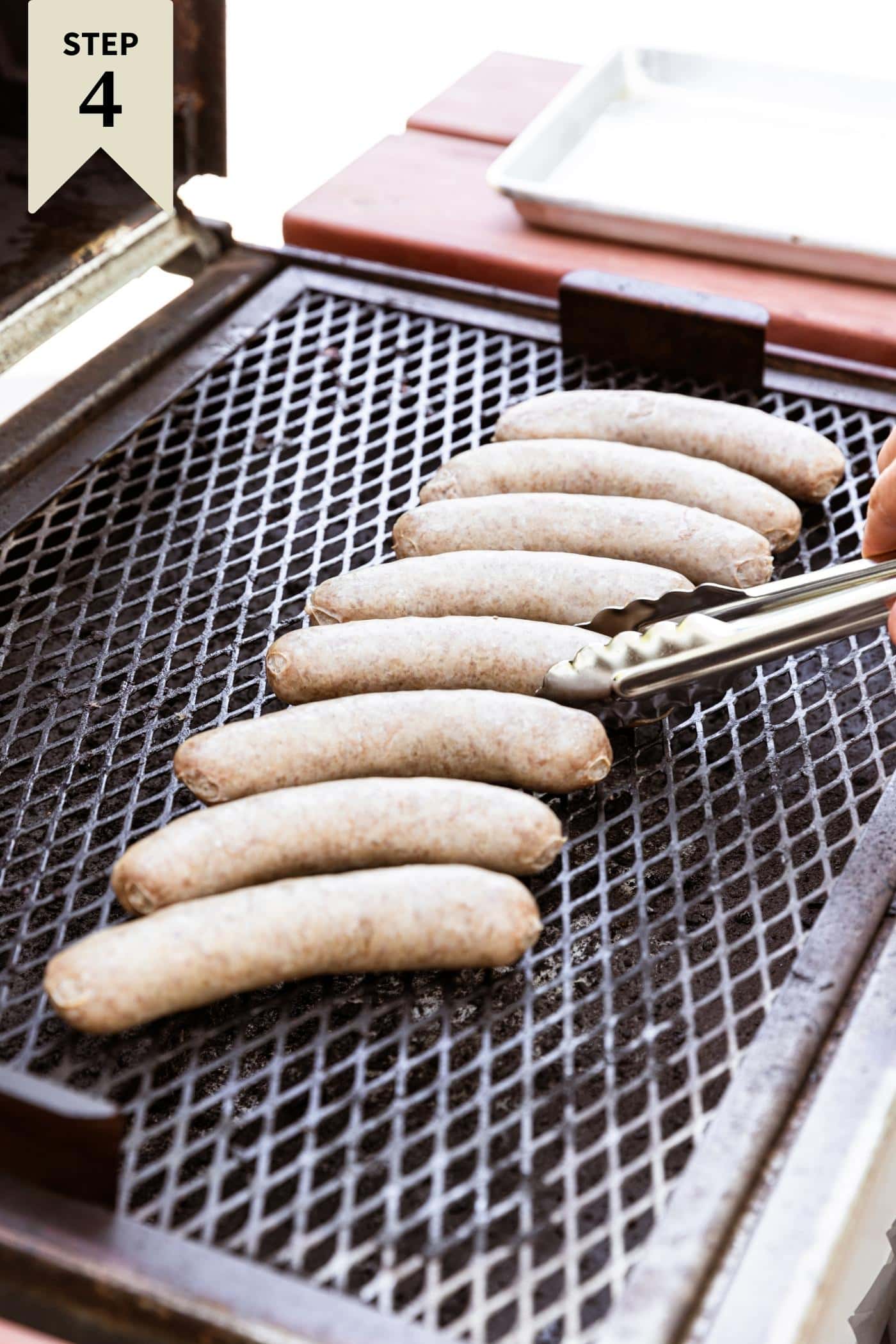 Grill with cover open with ungrilled brats sitting on grill top with tongs flipping one.