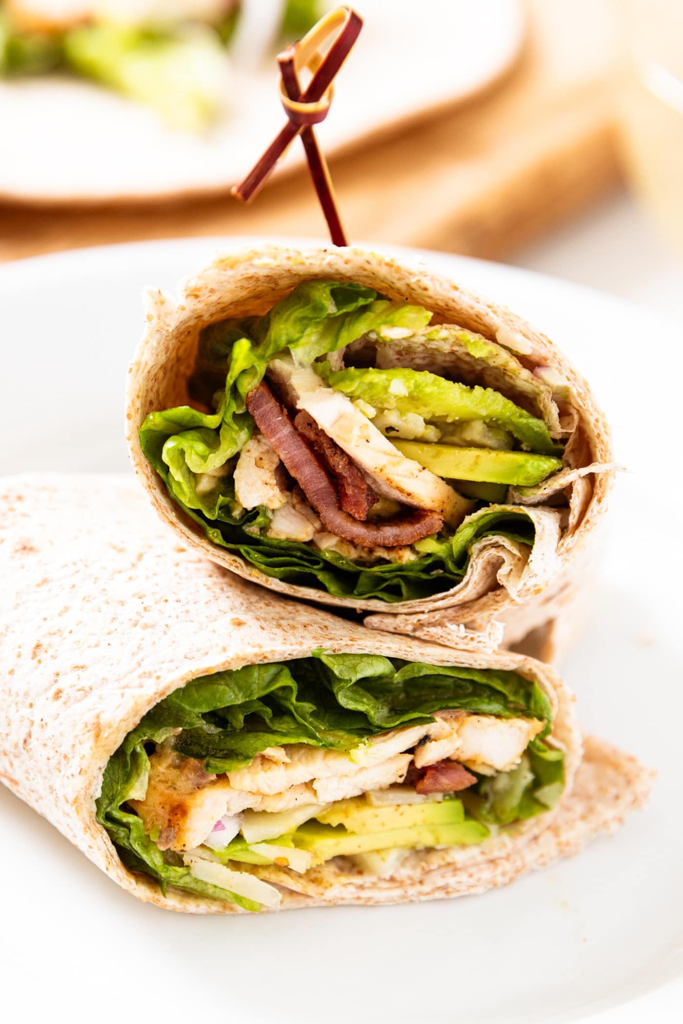 Chicken Caesar wrap cut in half sitting on a white plate showing the interior of the wrap filled with chicken, lettuce, avocado, bacon, and onion.