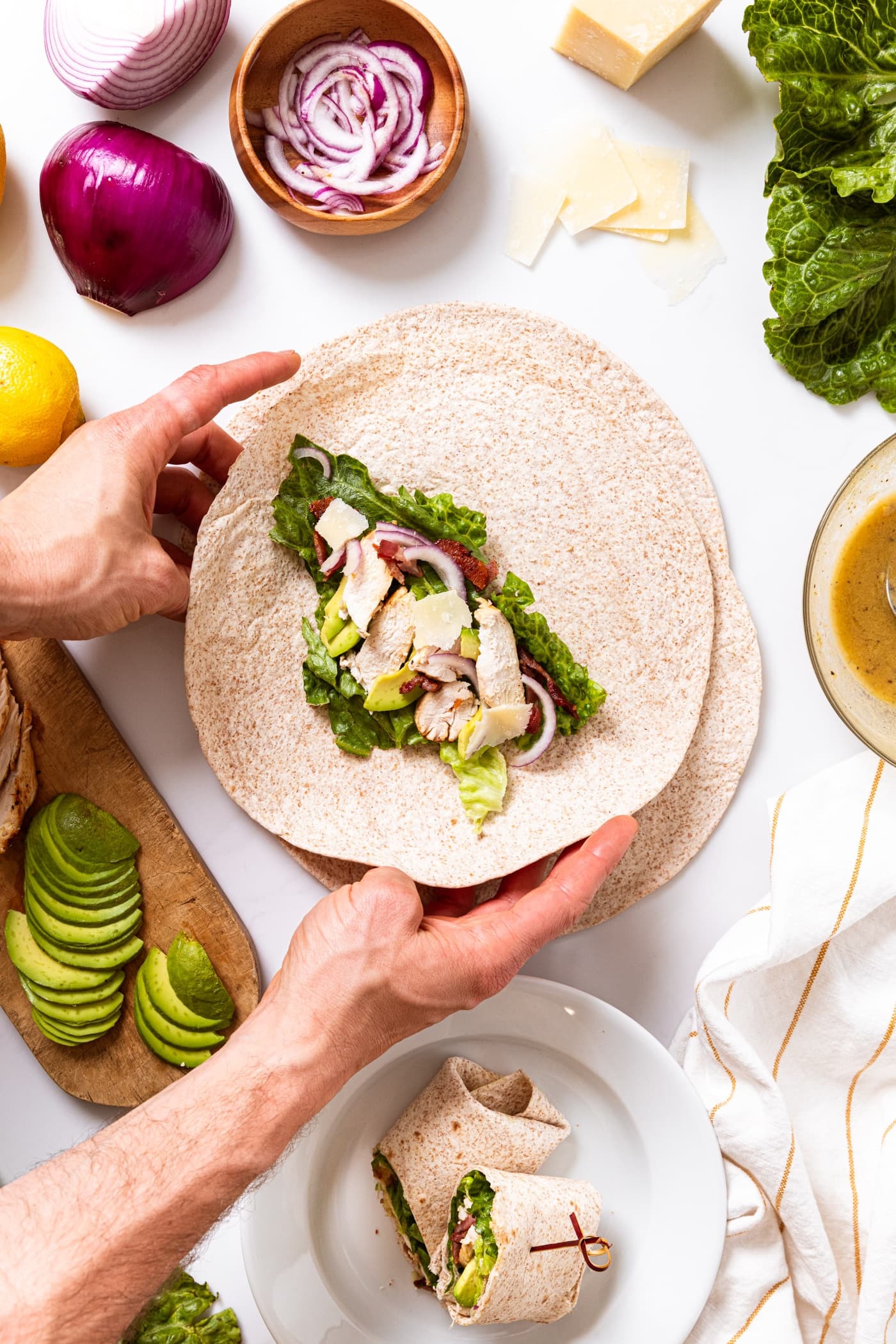 Two hands reaching to fold up flour tortilla with grilled chicken, onion, avocado, bacon, lettuce, and cheese on top.
