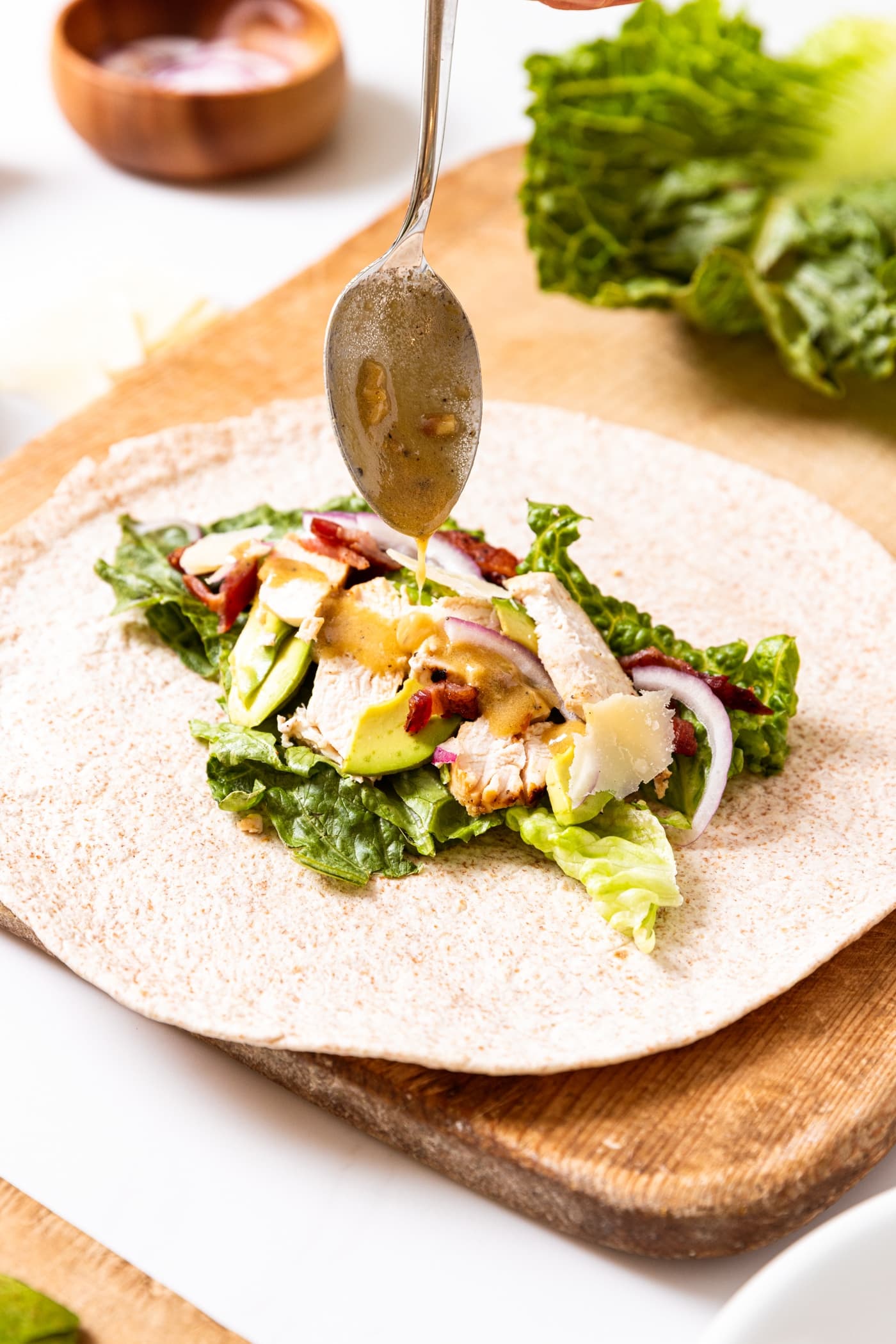 Open faced flour tortilla with strips of chicken, lettuce, bacon, avocado, and Parmesan cheese with spoon drizzling on Caesar dressing.