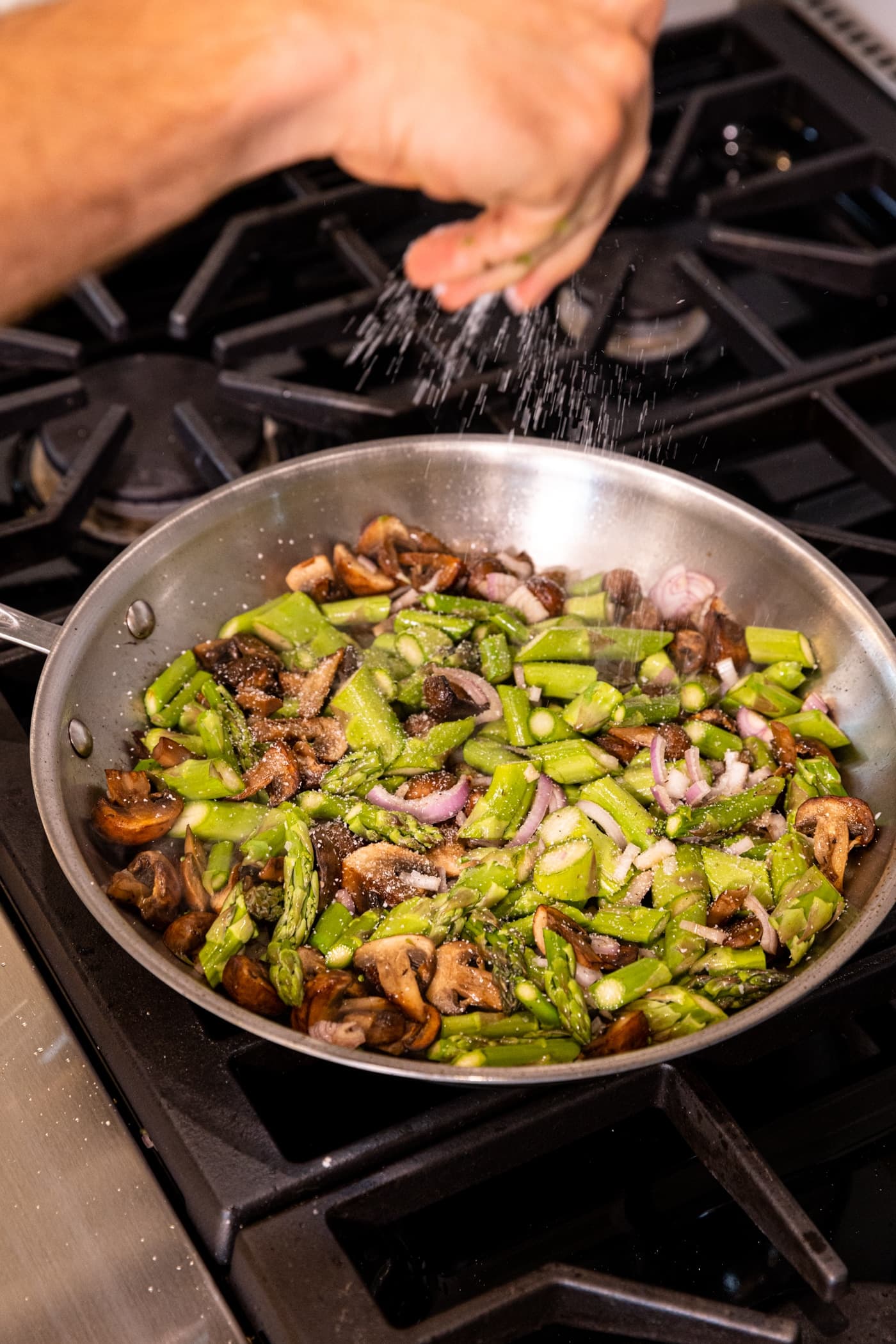 Skillet on stove with pieces of asparagus and brown mushrooms with hand sprinkled salt over the top.