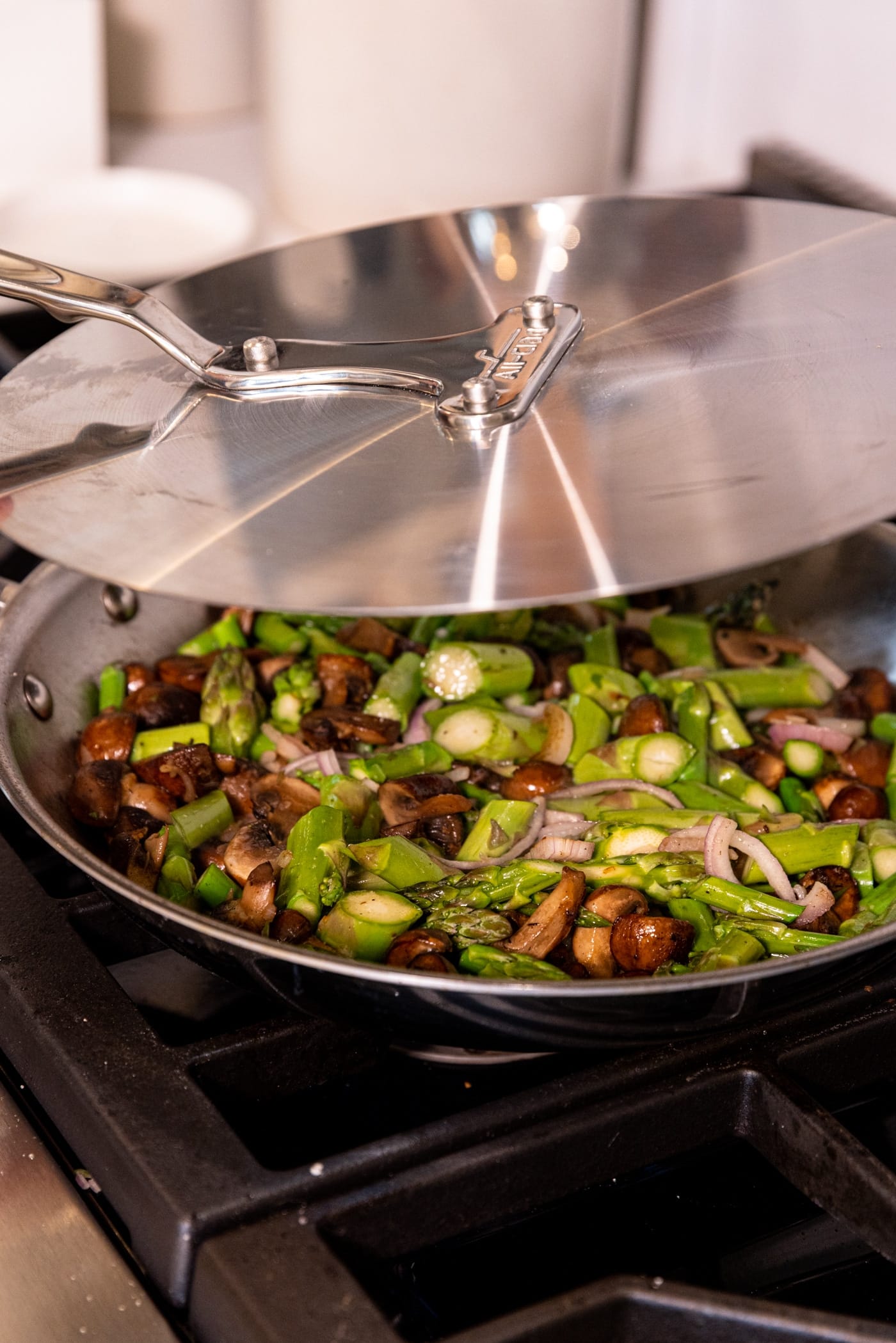 Stainless steel skillet on stove filled with pieces of asparagus and mushroom pieces with lid being placed on top.
