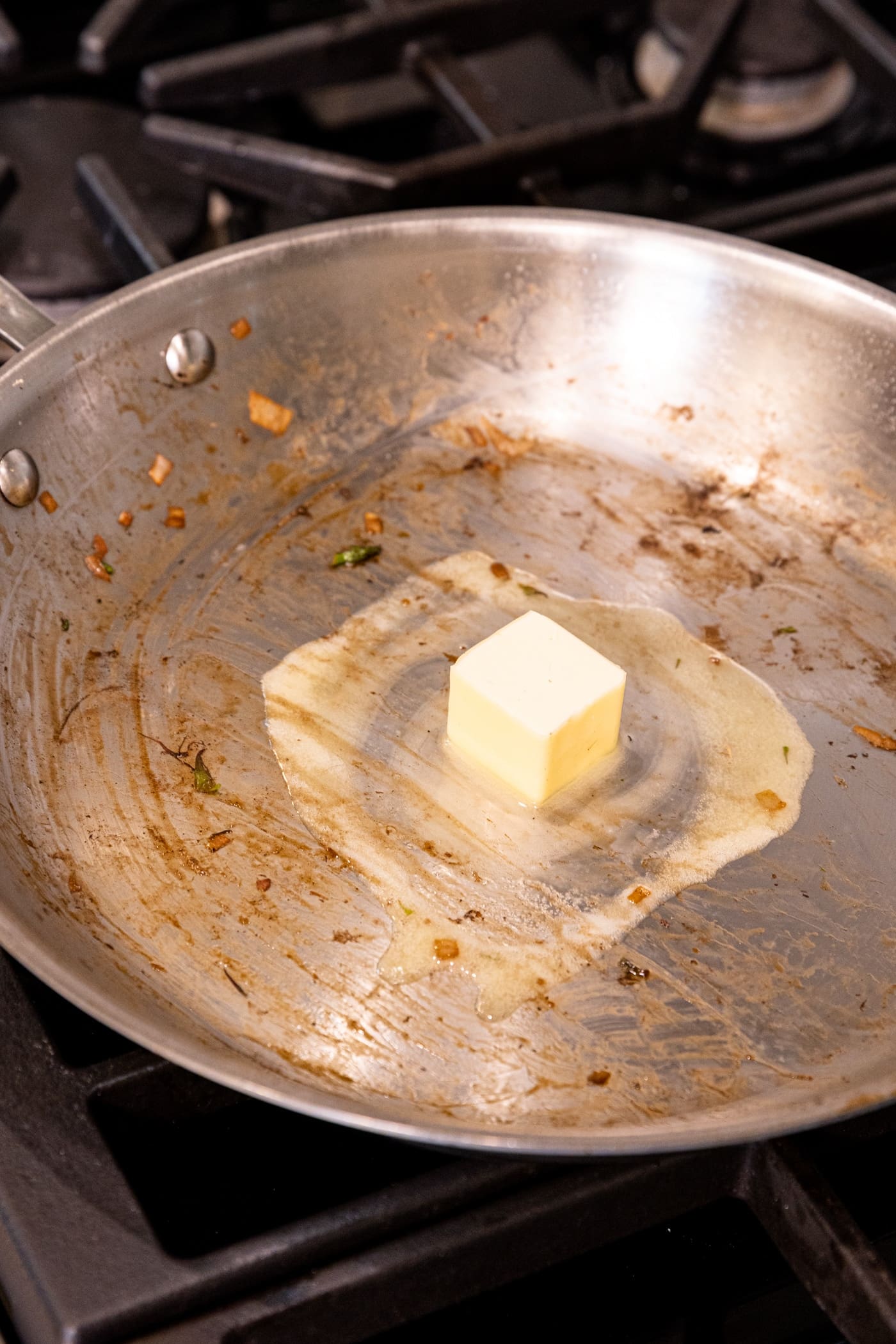 Square of light colored butter melting in skillet with debris from previous cooking.