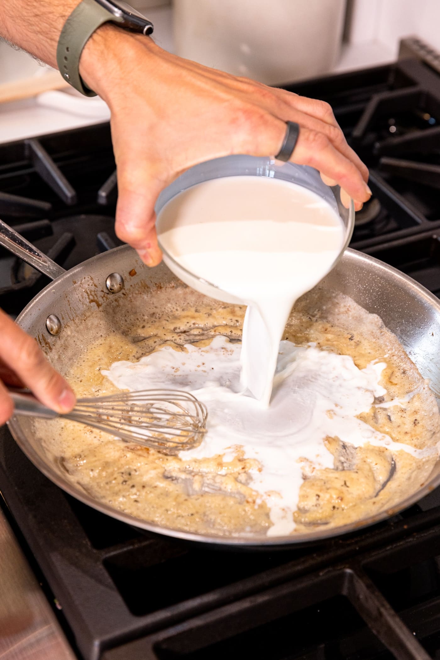 Hand pouring small glass bowl filled with milk into skillet on stove with roux mixture.