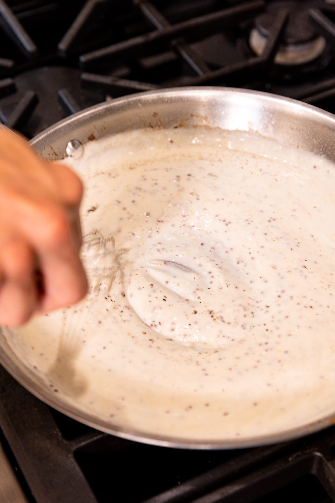 Hand stirring white sauce with whisk in skillet on stovetop.