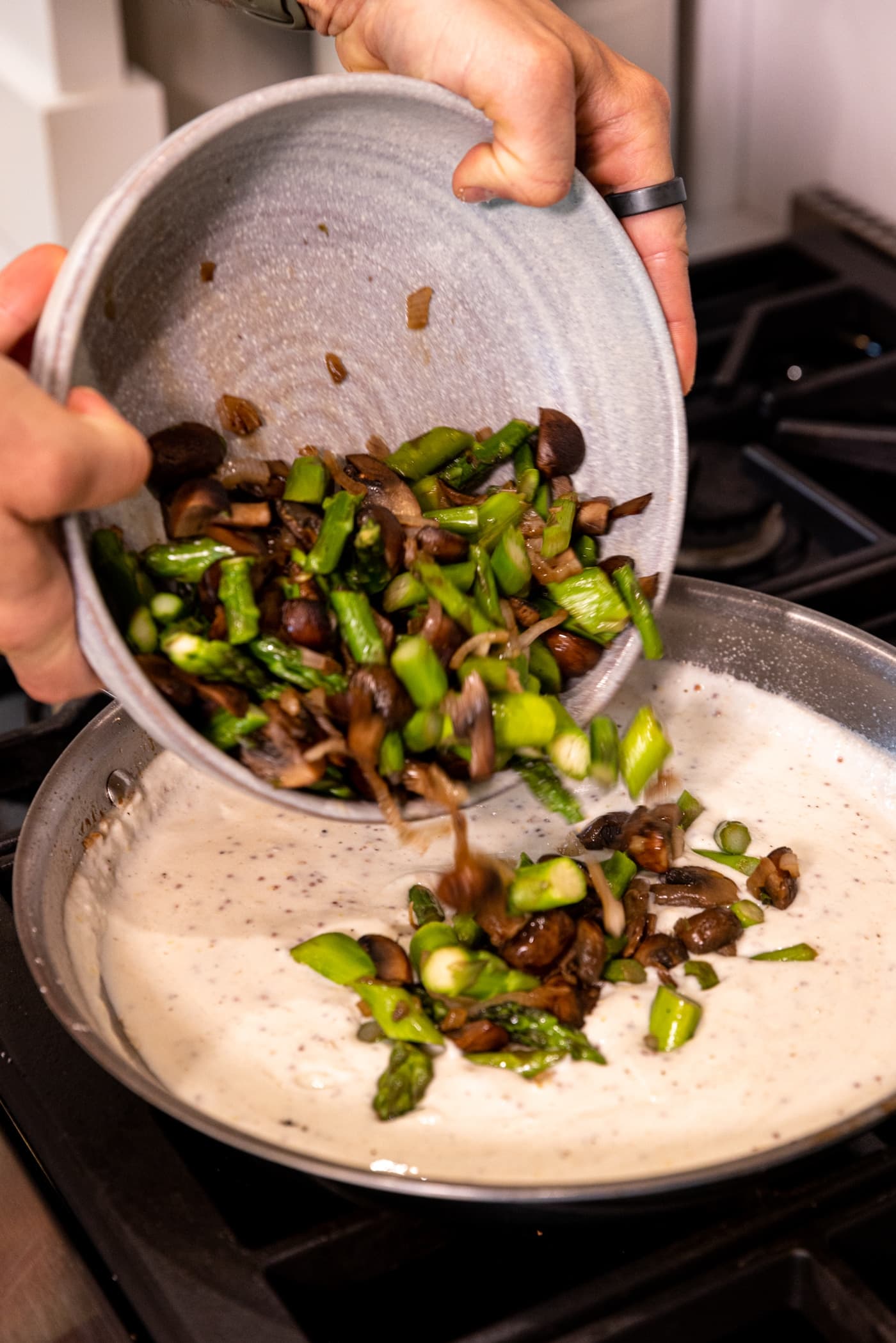 Stoneware bowl filled with asparagus and mushrooms being poured into white sauce in skillet on stovetop.