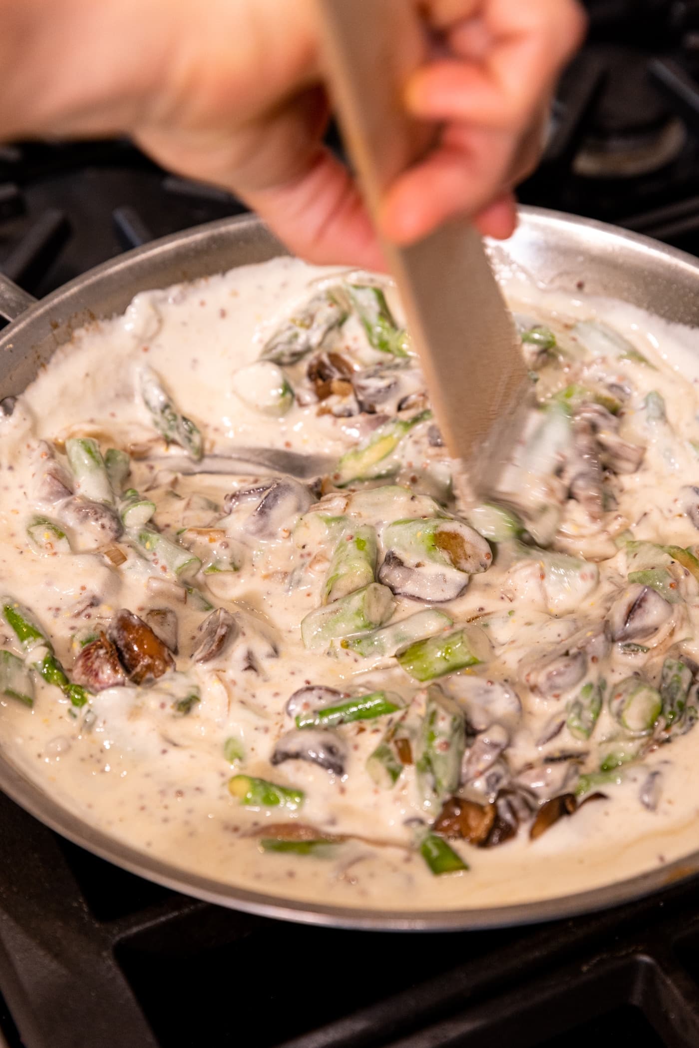 Wooden spatula stirring in asparagus and mushrooms into white sauce in skillet.