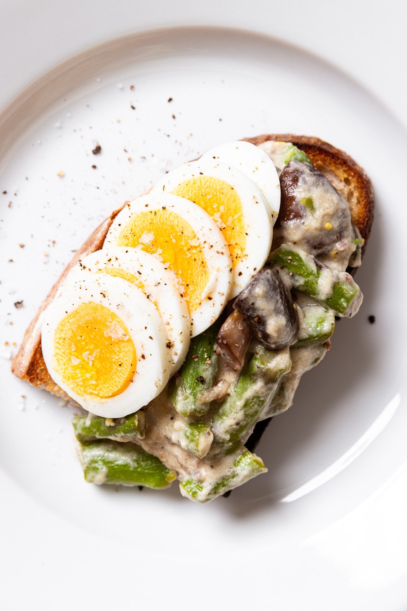 White plate filled with half piece of toast with creamed asparagus and hardboiled egg on top.