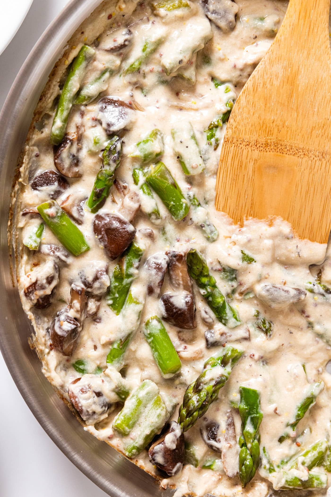 Stainless steel skillet filled with mushroom and asparagus mixture sitting in white colored cream sauce with wooden spatula in pan as well.