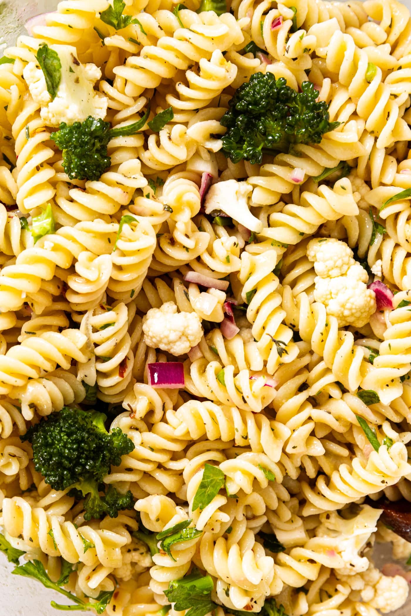 Light yellow colored spiral pasta noodles mixed with pieces of broccoli, cauliflower, and red onion with Italian dressing poured over top in glass bowl.