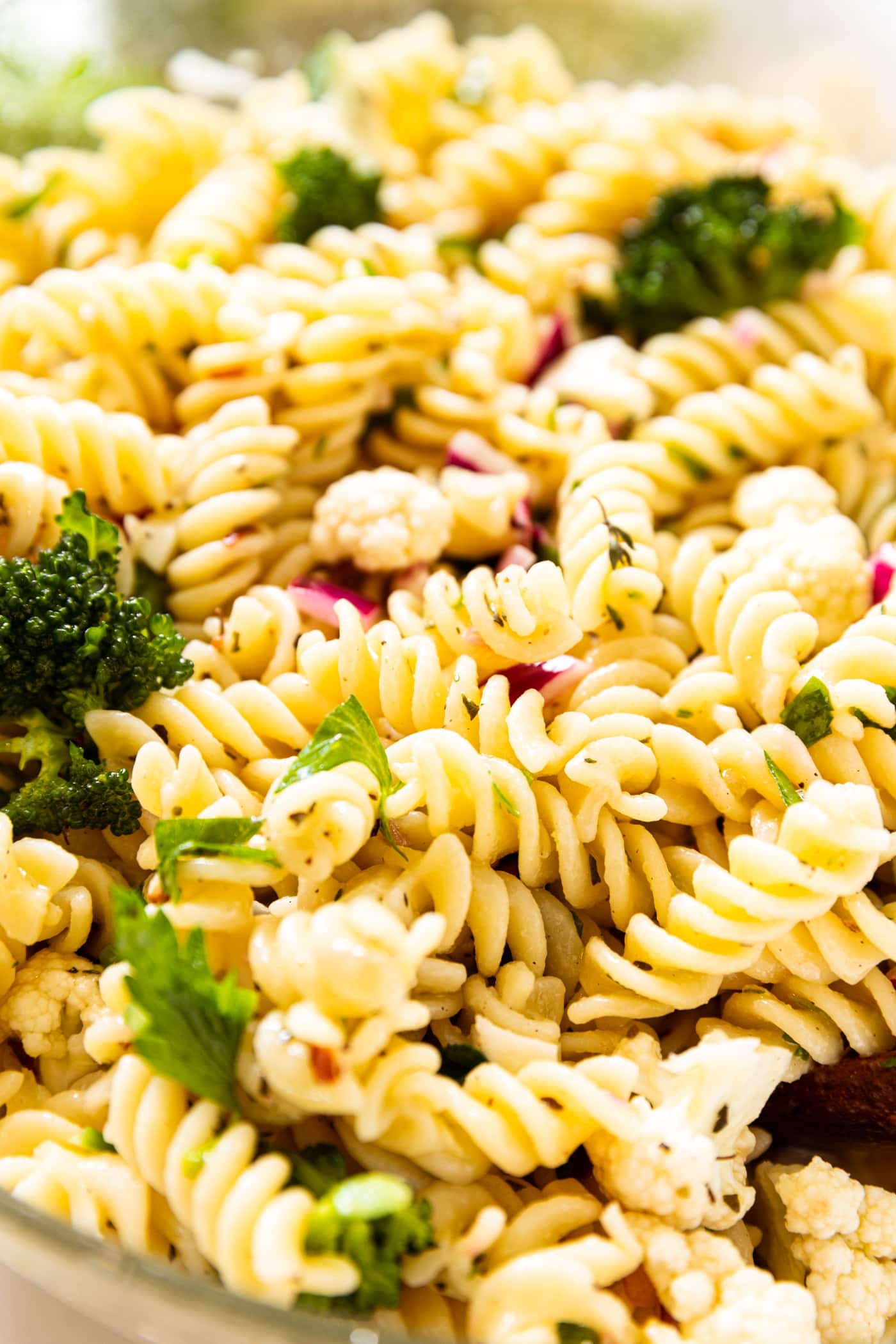 Glass bowl filled with yellow colored spiral pasta with pieces of broccoli, cauliflower, red onion, and chopped parsley mixed in.