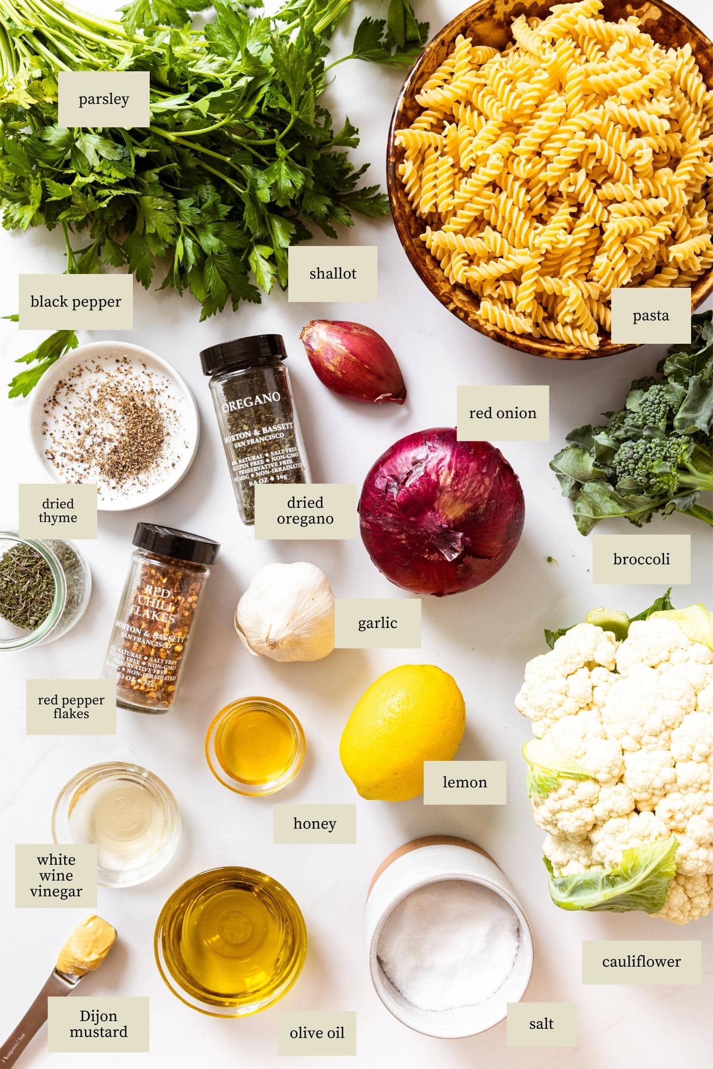 White countertop filled with all types of ingredients needed to make pasta salad recipe including big bowl of pasta, red onion, spices, cauliflower, and broccoli.