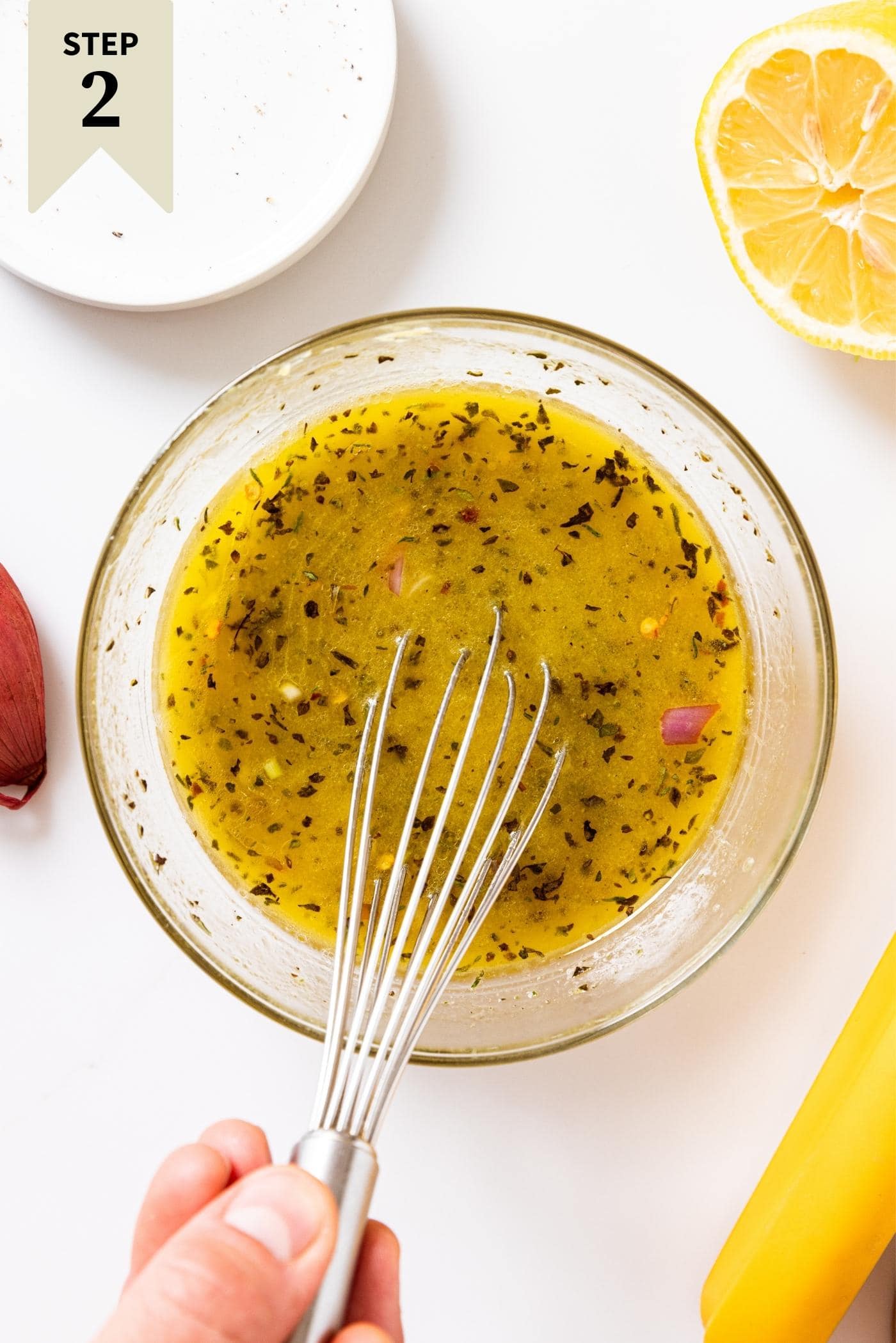 Clear glass bowl sitting on white countertop filled with Italian dressing with hand whisking contents of bowl.