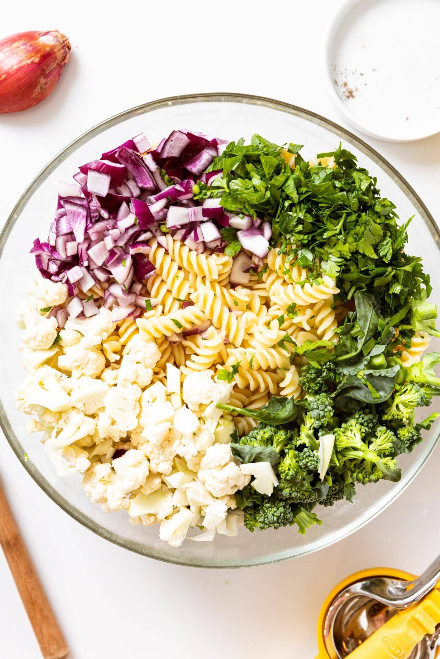Large glass bowl filled with pasta with red onion, parsley, broccoli, and cauliflower all around the circumference of the bowl.