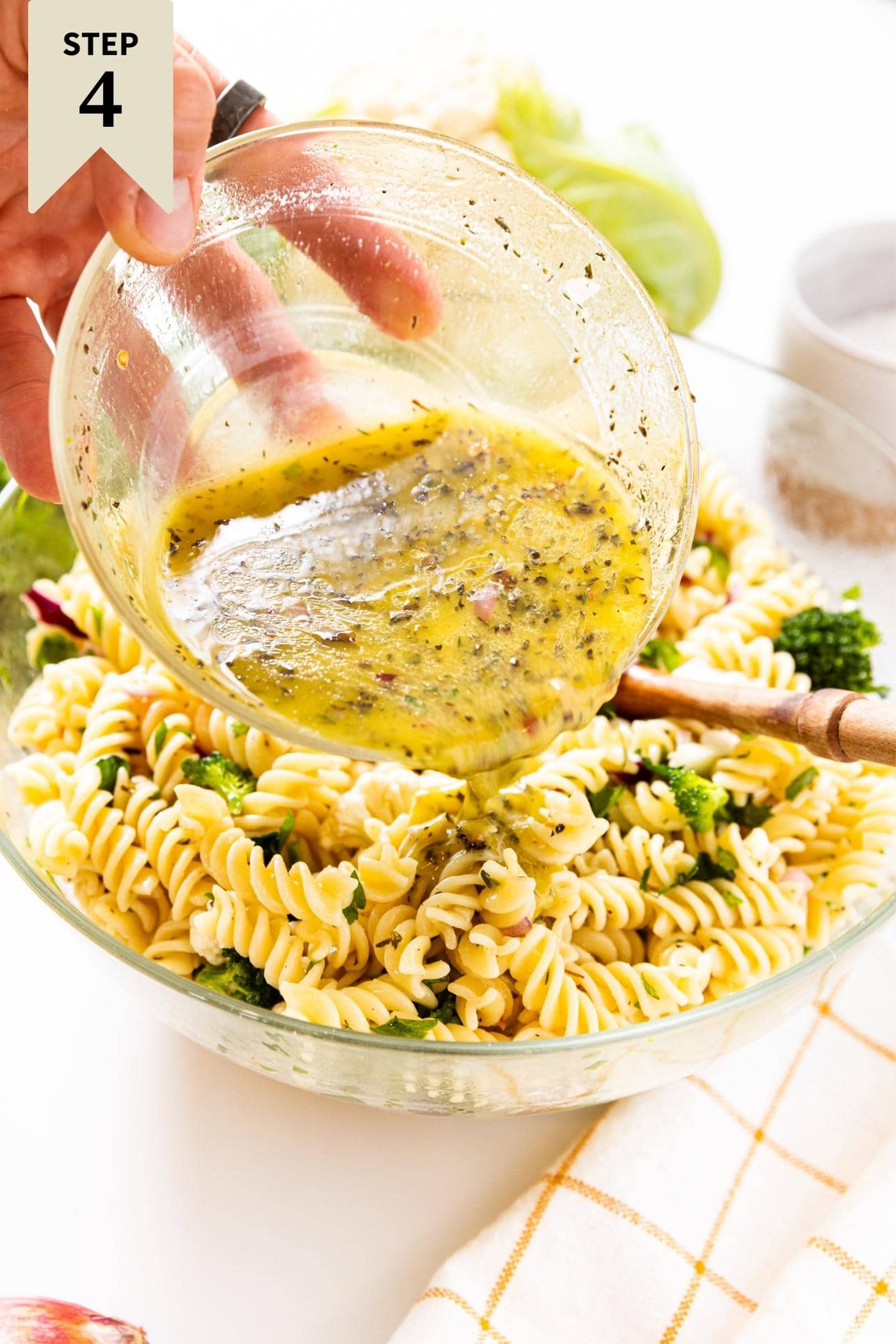 Hand holding glass bowl pouring Italian dressing on top of mixed pasta salad with spiral pasta noodles.