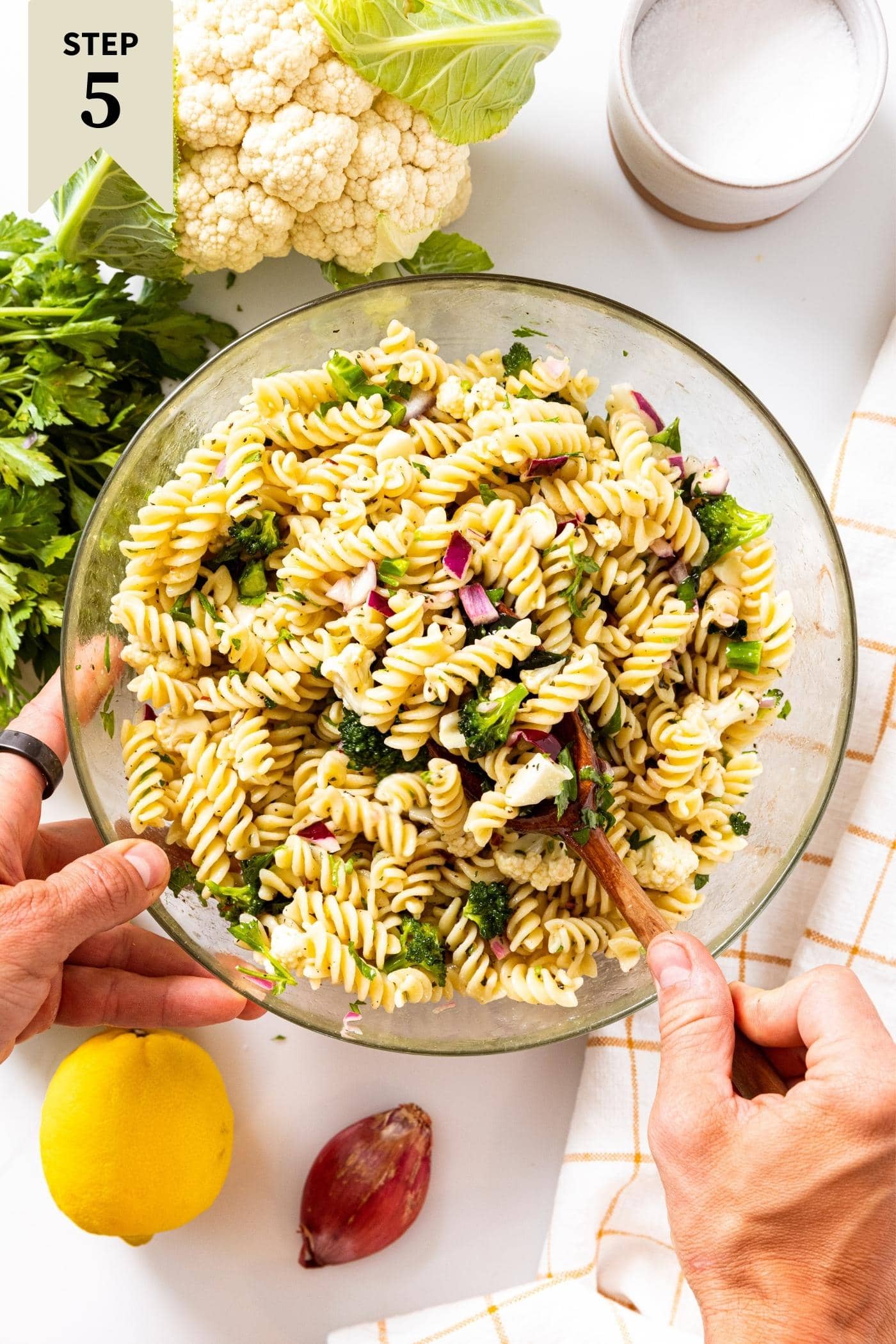 Hand holding wooden spoon stirring pasta salad with spiral pasta and broccoli and cauliflower.