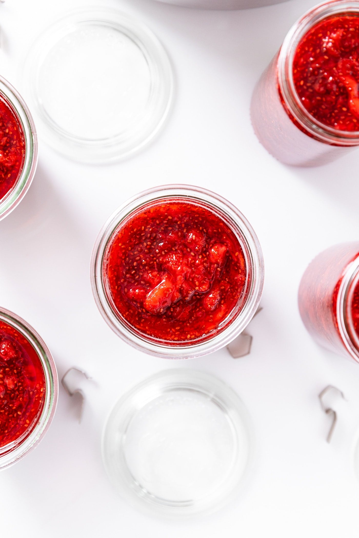 Glass container filled with red colored strawberry jam with chia seeds showing with glass lids sitting on marble countertop along with other glass jars.