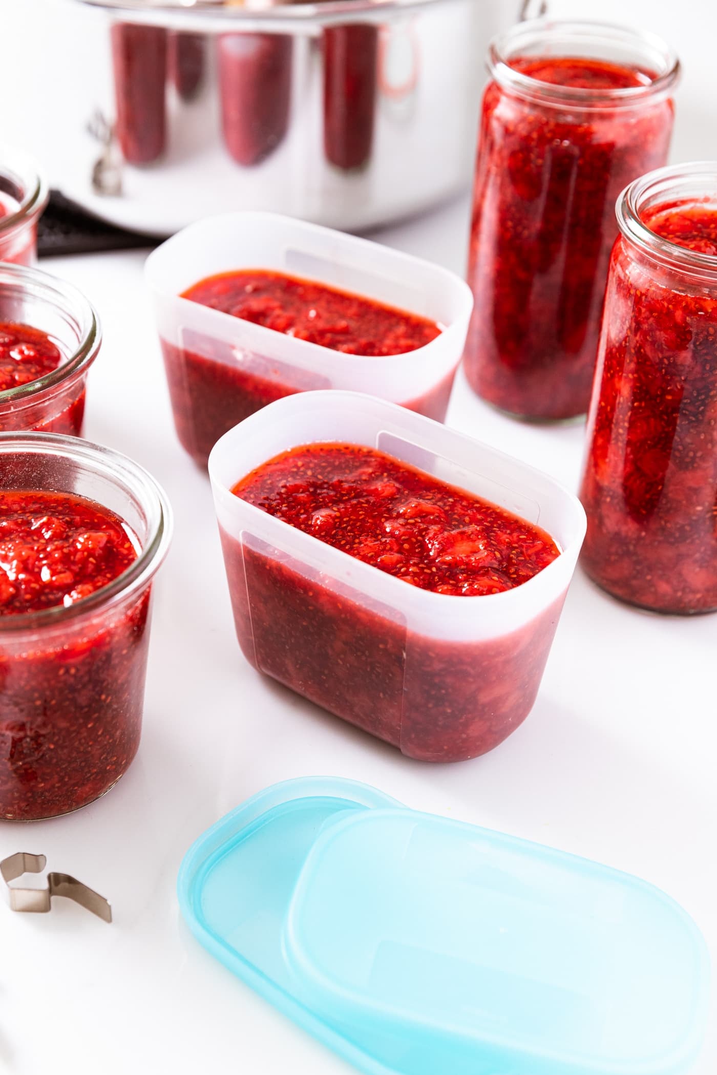 Plastic containers filled with strawberry freezer jam with blue lids sitting in front with glass jars sitting around also filled with red colored jam.