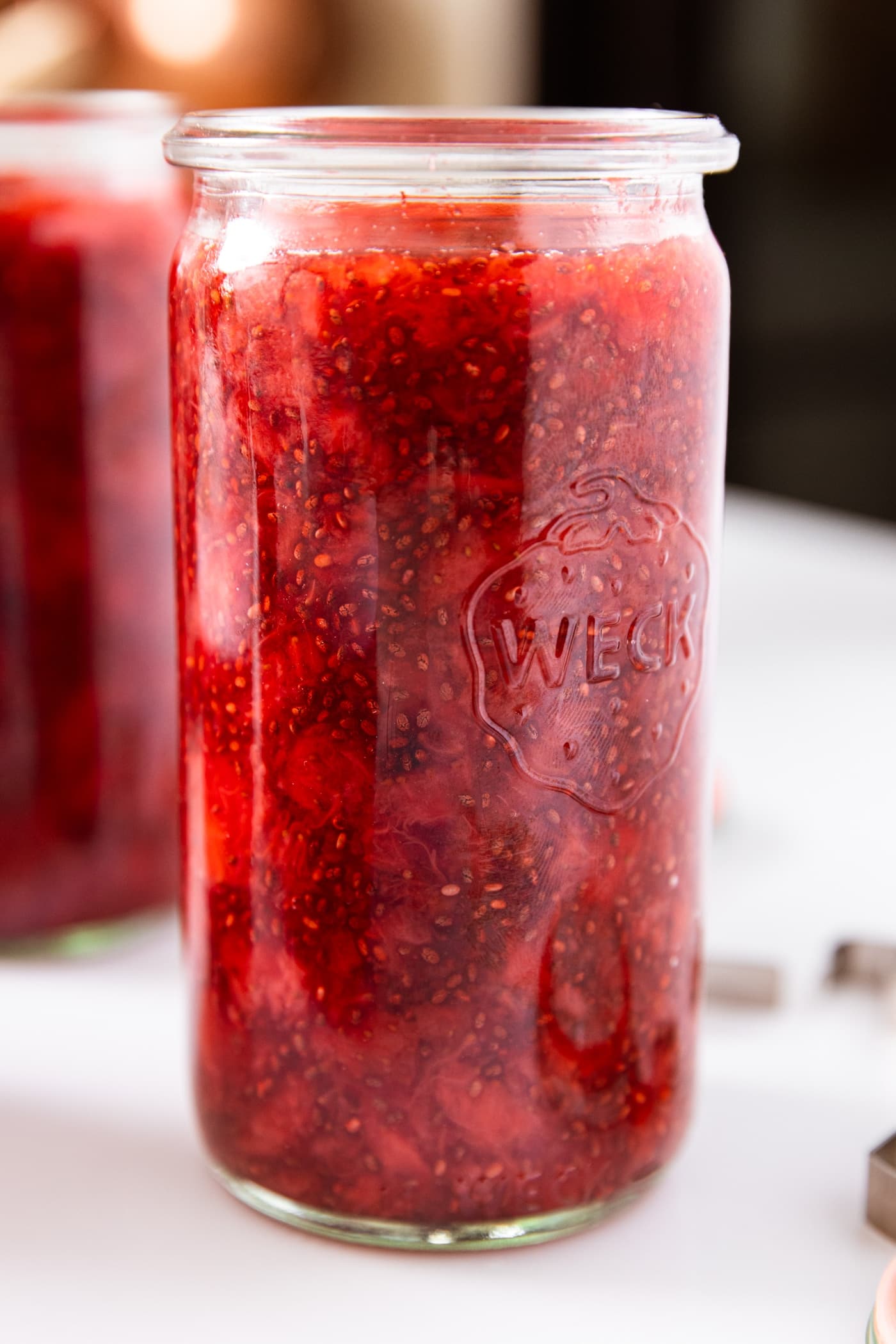 Large tall glass jar filled with strawberry freezer jam with chia seeds showing sitting on white surface with more jars in background.