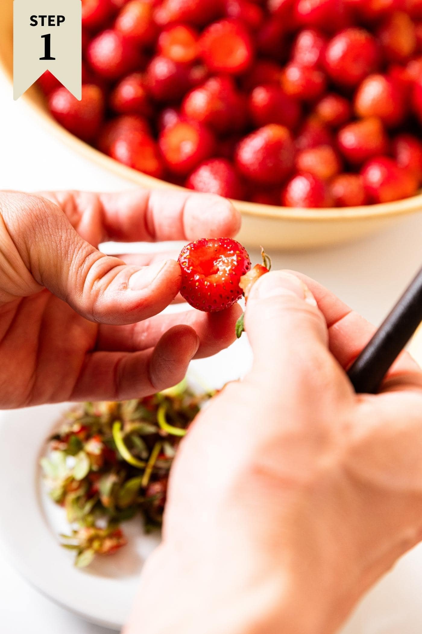 Two hands holding hulled strawberry with knife also in hand with discarded stems below and more hulled strawberries in background.