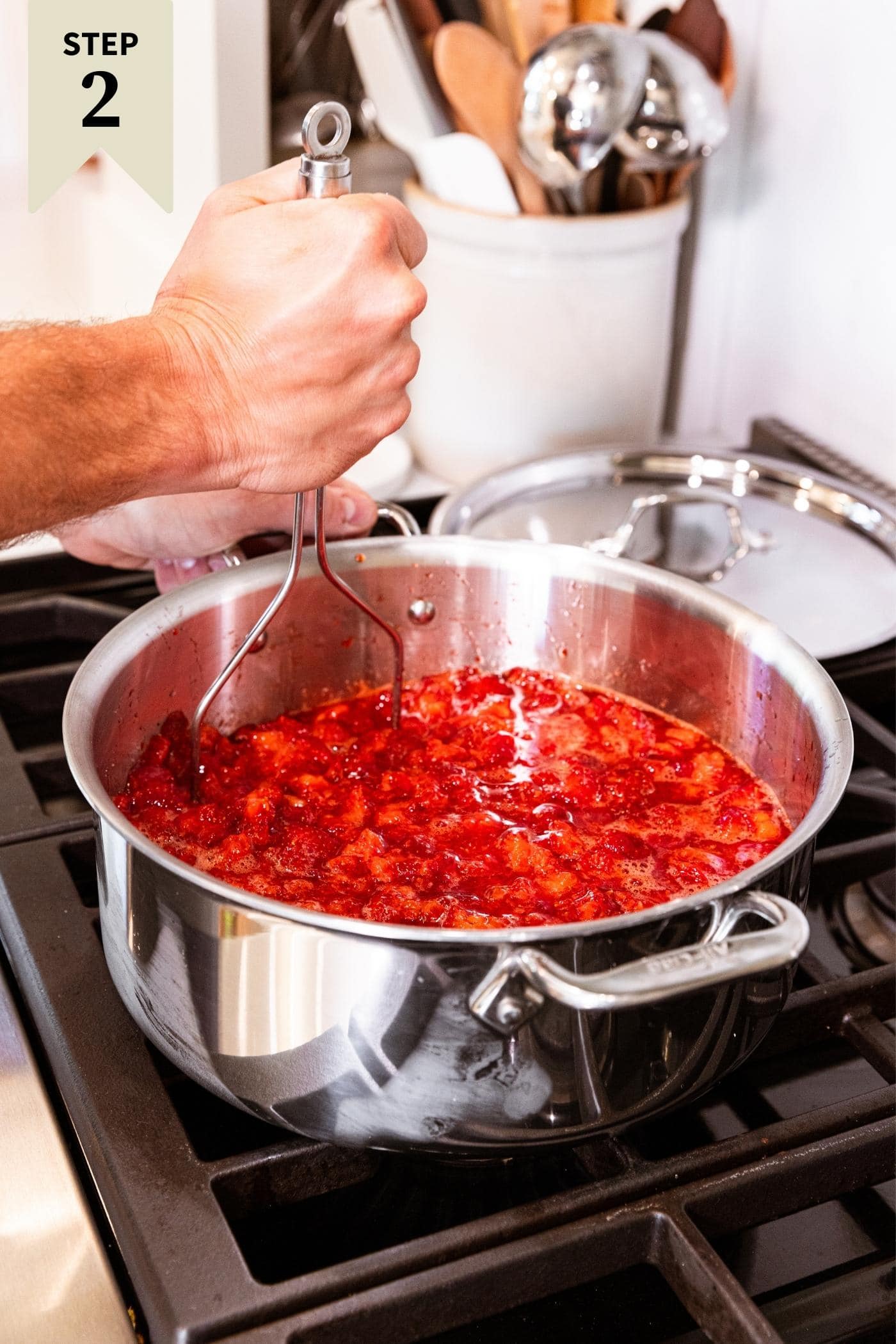 Hand holding potato masher smashing strawberries in metal pot on stovetop with metal lid in background.