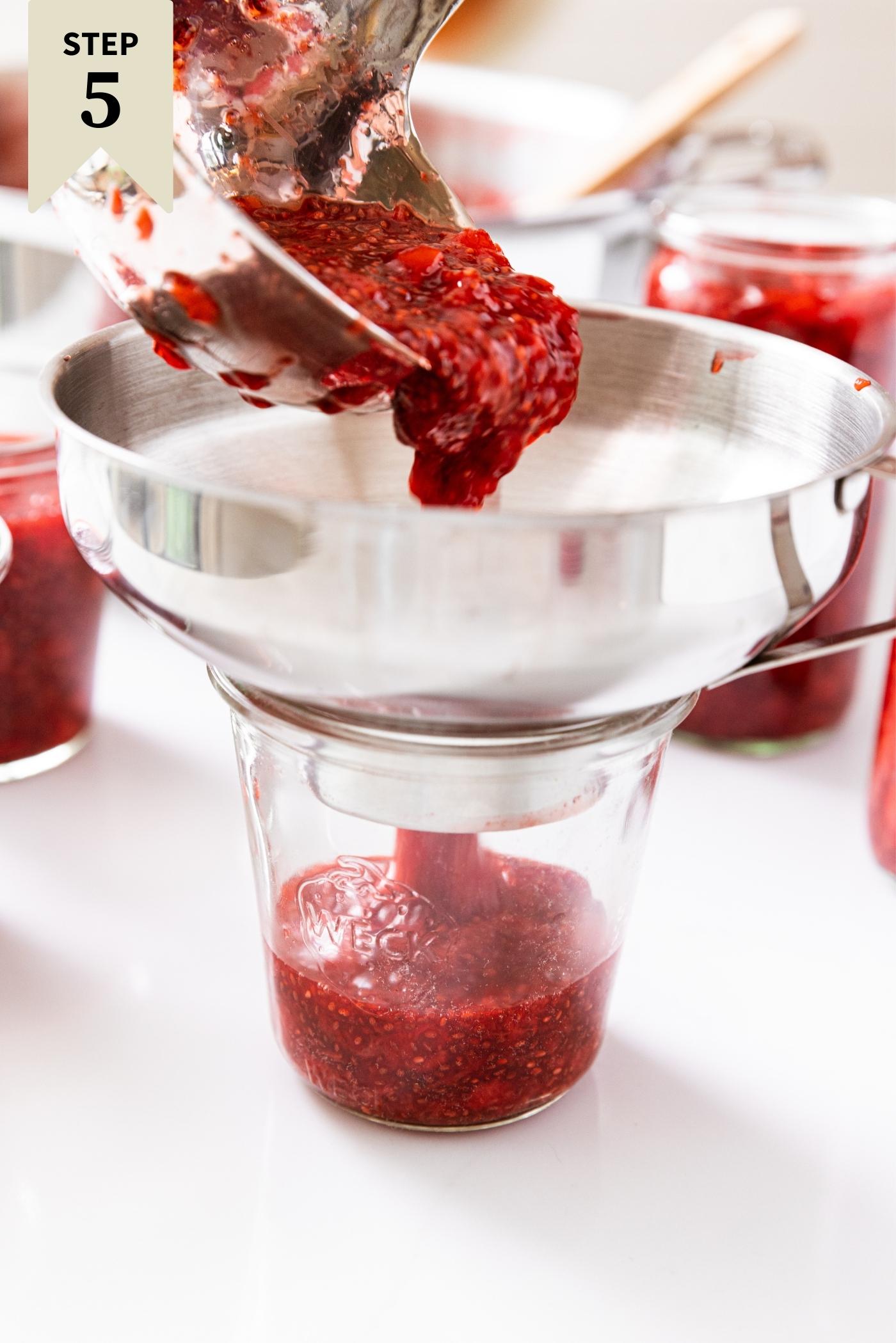 Metal funnel fitted on top of glass jar with strawberry jam being ladled into jar.