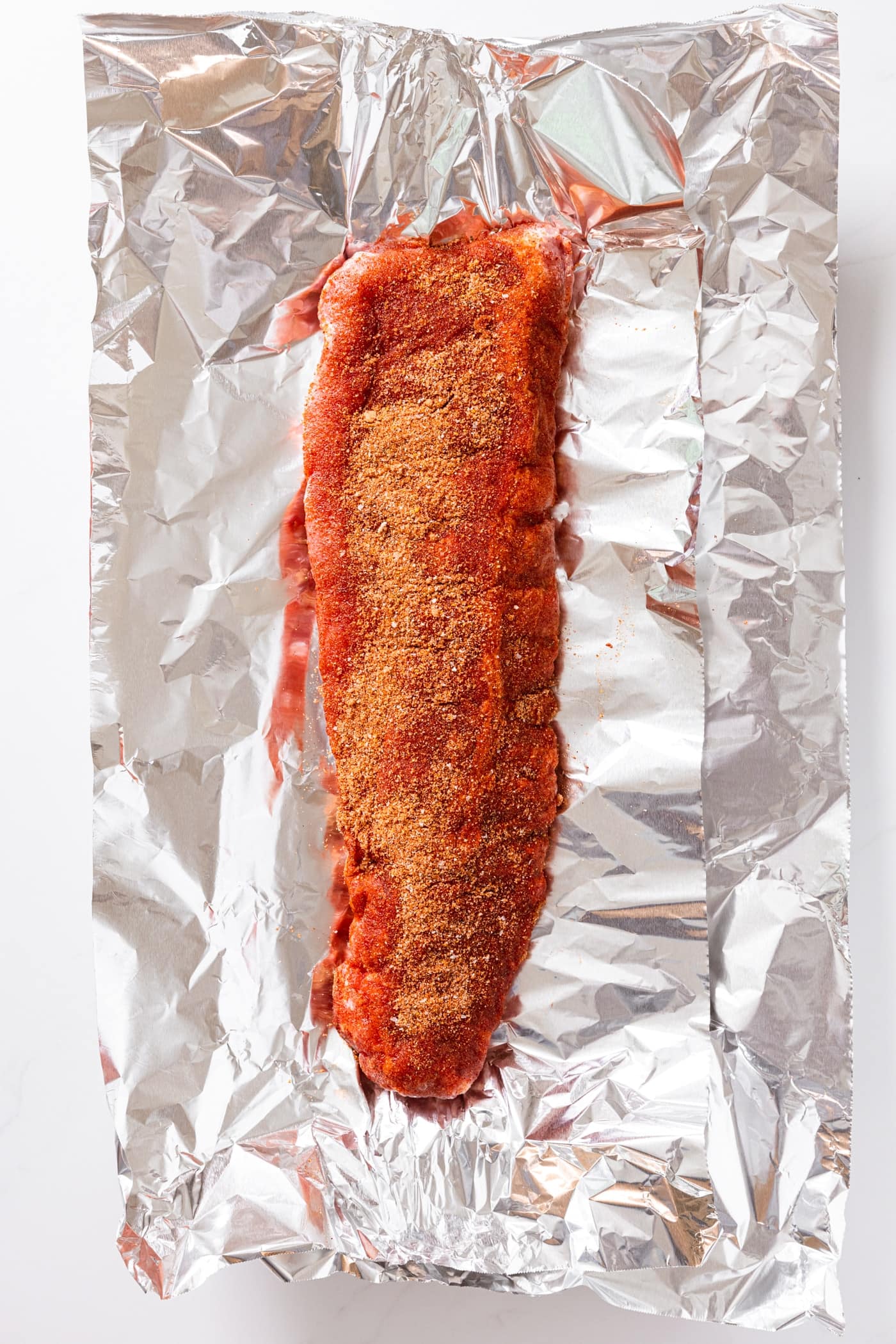 Two pieces of tin foil sitting on white countertop with rack of baby back ribs with spice rub on top sitting on foil.