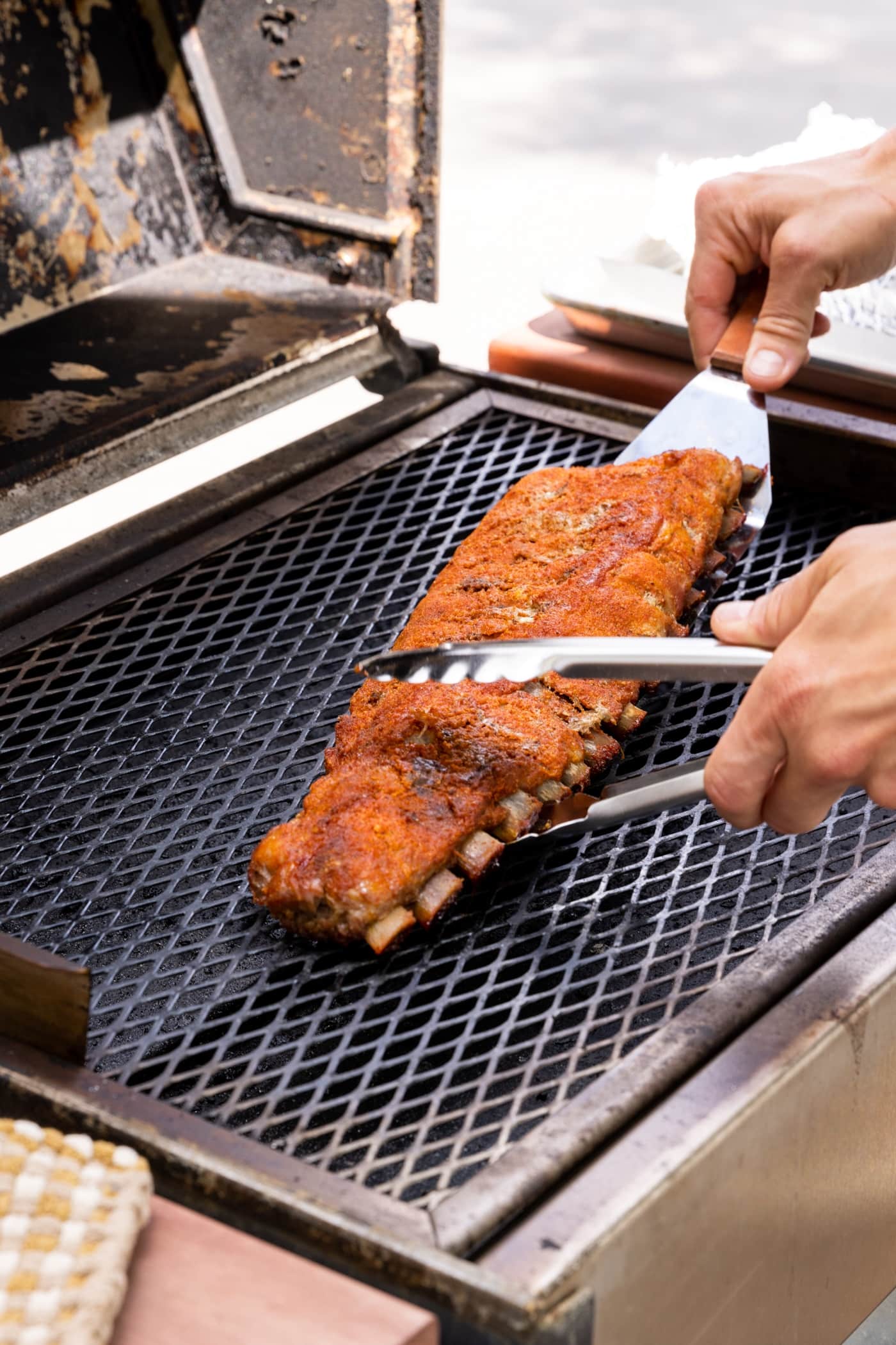 Two hands using tongs to place an uncooked rack of ribs on grill surface.
